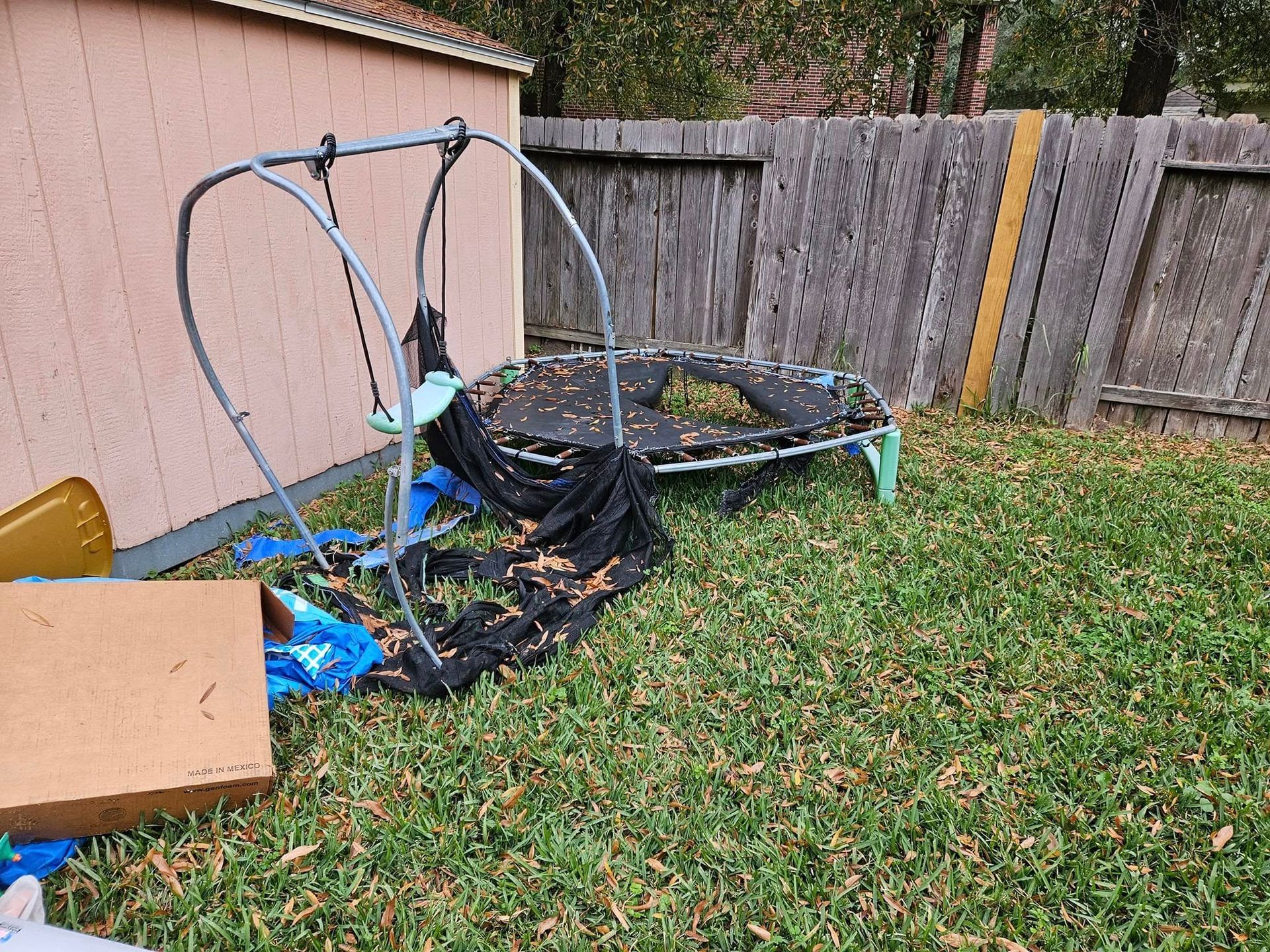 Broken trampoline and scattered debris in a grassy backyard beside a shed and wooden fence