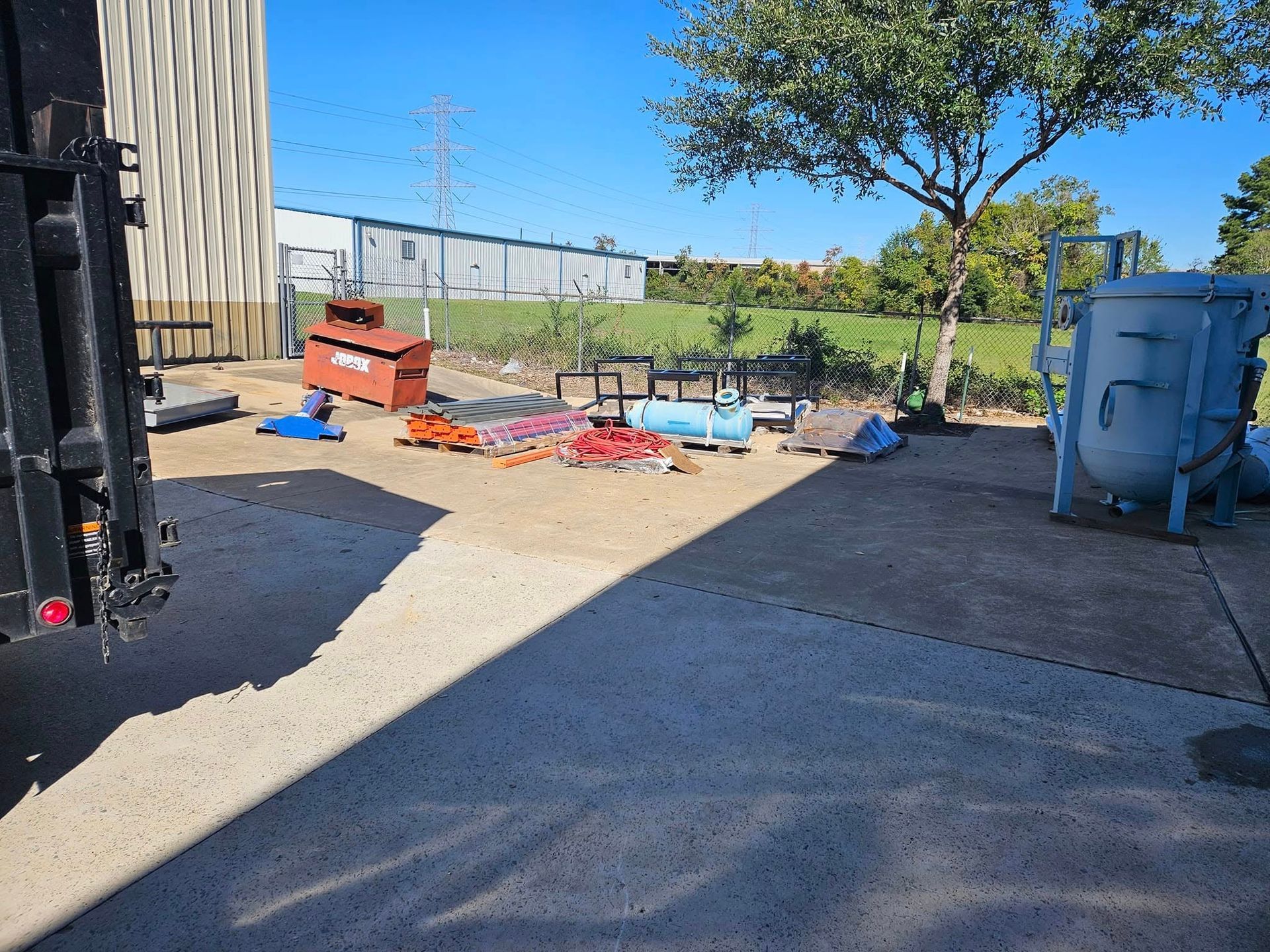 Outdoor construction yard with scattered debris, a blue bin, and a fenced area under bright sunlight