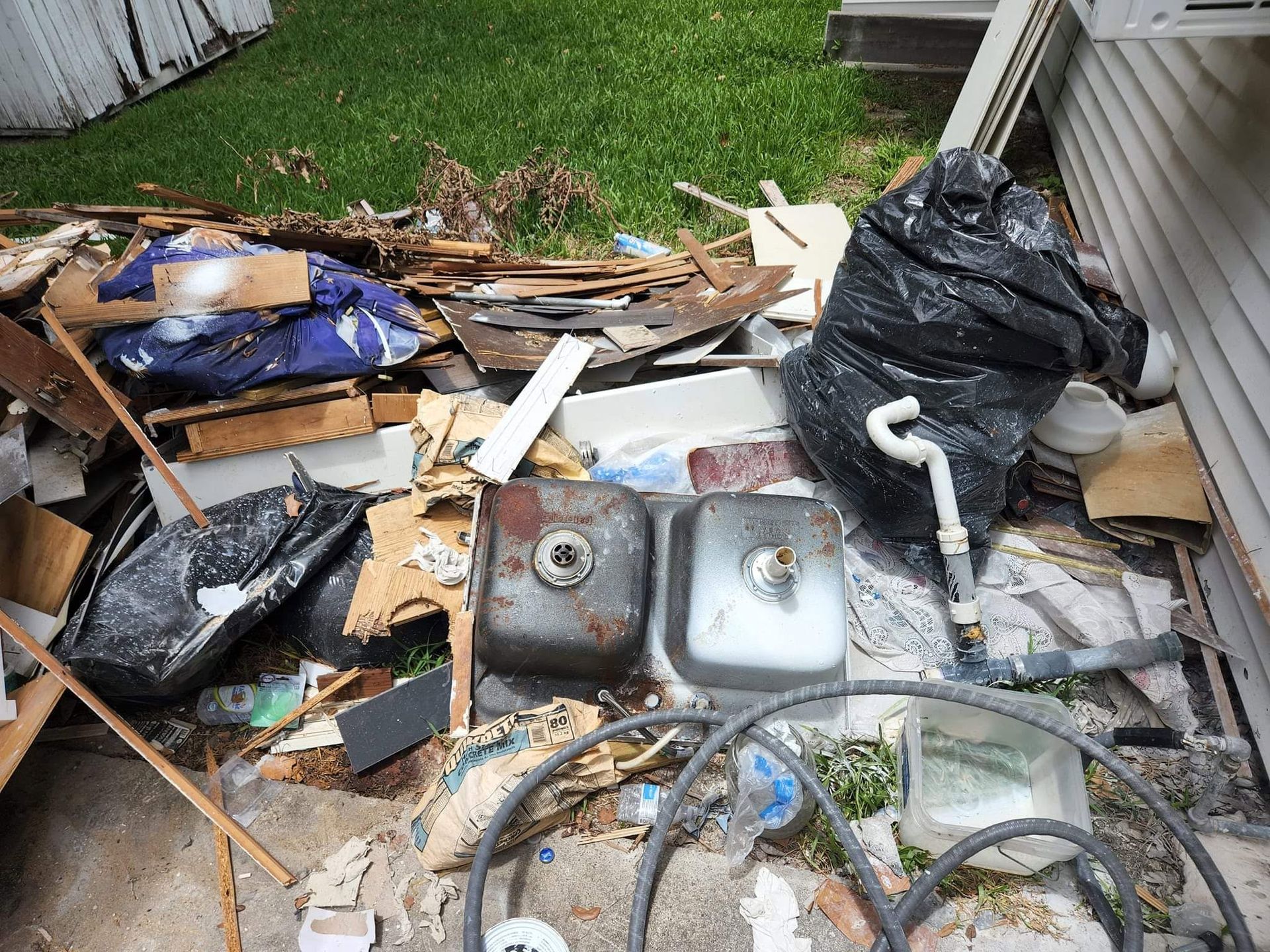 Piles of debris and broken household items scattered along a house exterior near a grassy yard.
