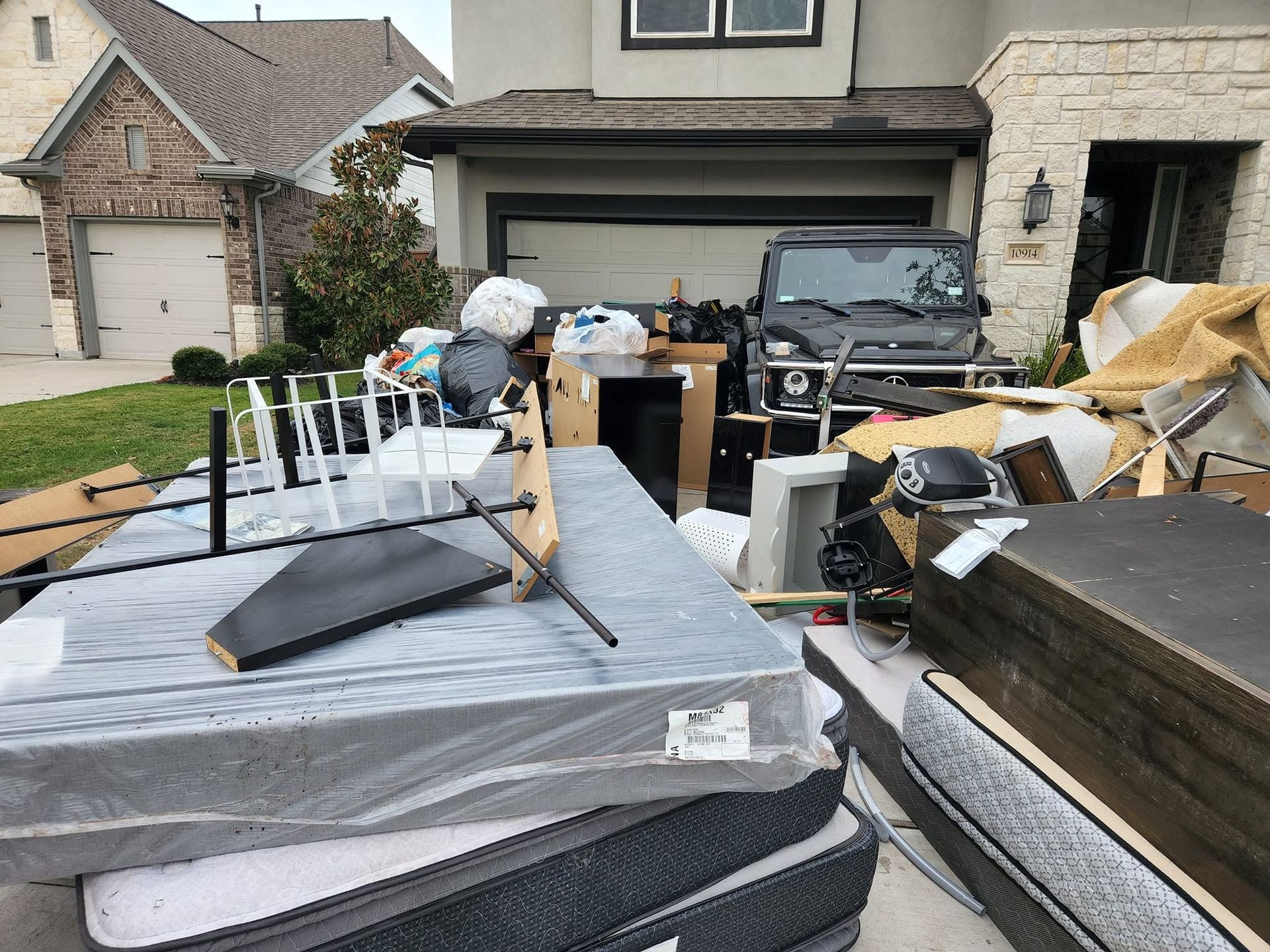 Front yard cluttered with stacked mattresses, furniture, and trash in front of a house and garage.