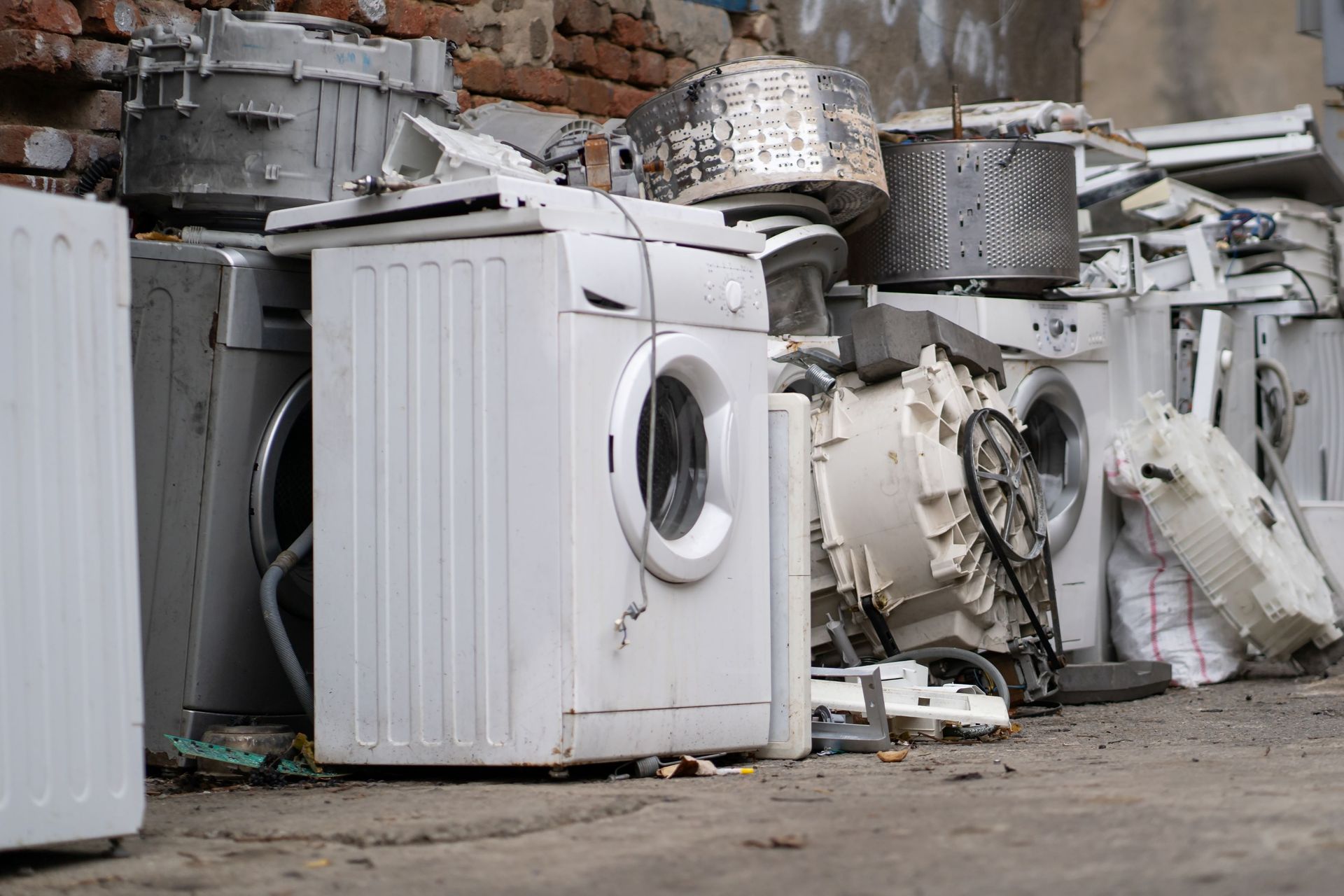Pile of old washing machines and metal scrap in an outdoor junkyard against a brick wall.