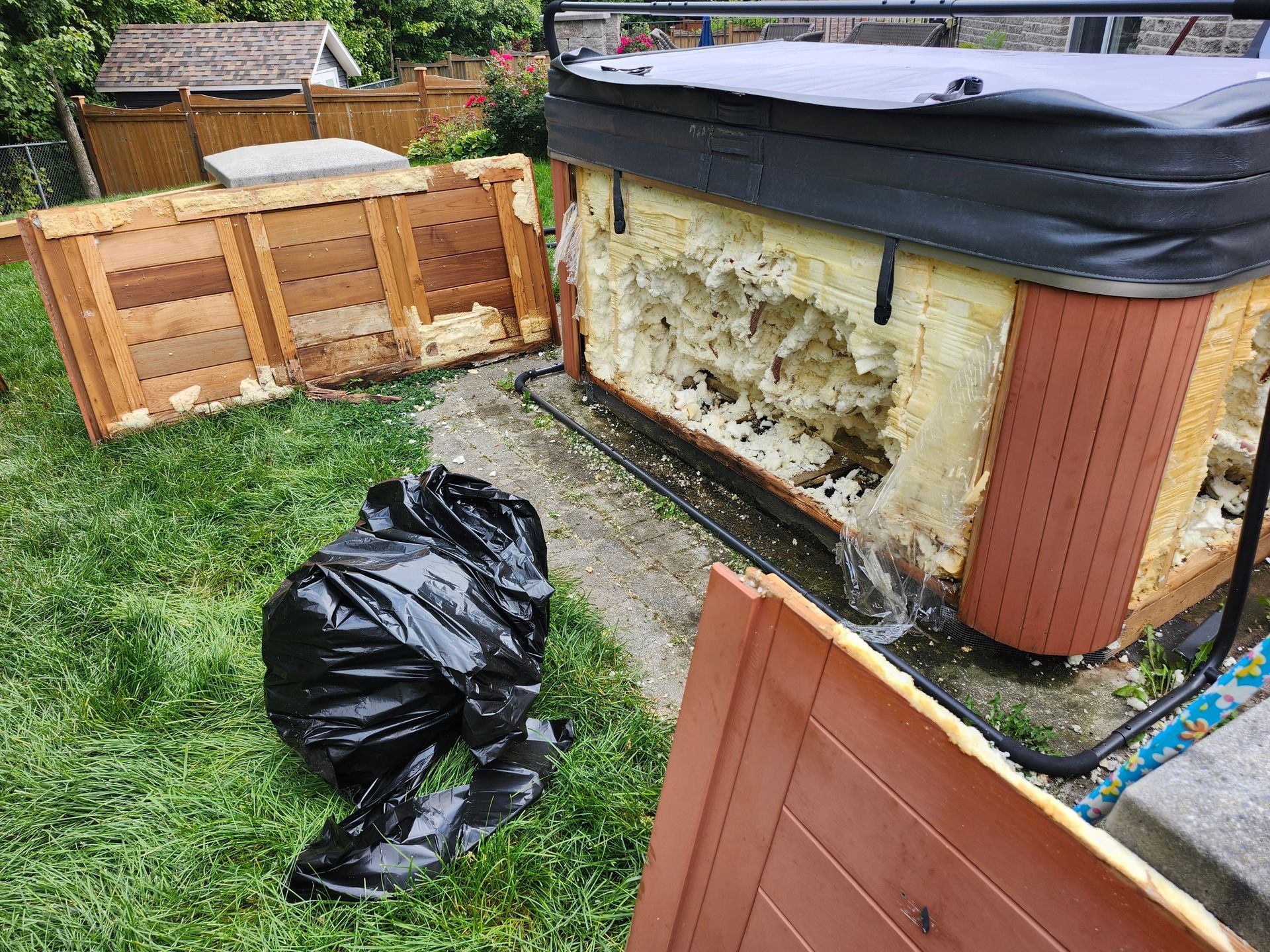 Damaged hot tub with foam insulation exposed, broken wooden panels, and black trash bags on grass