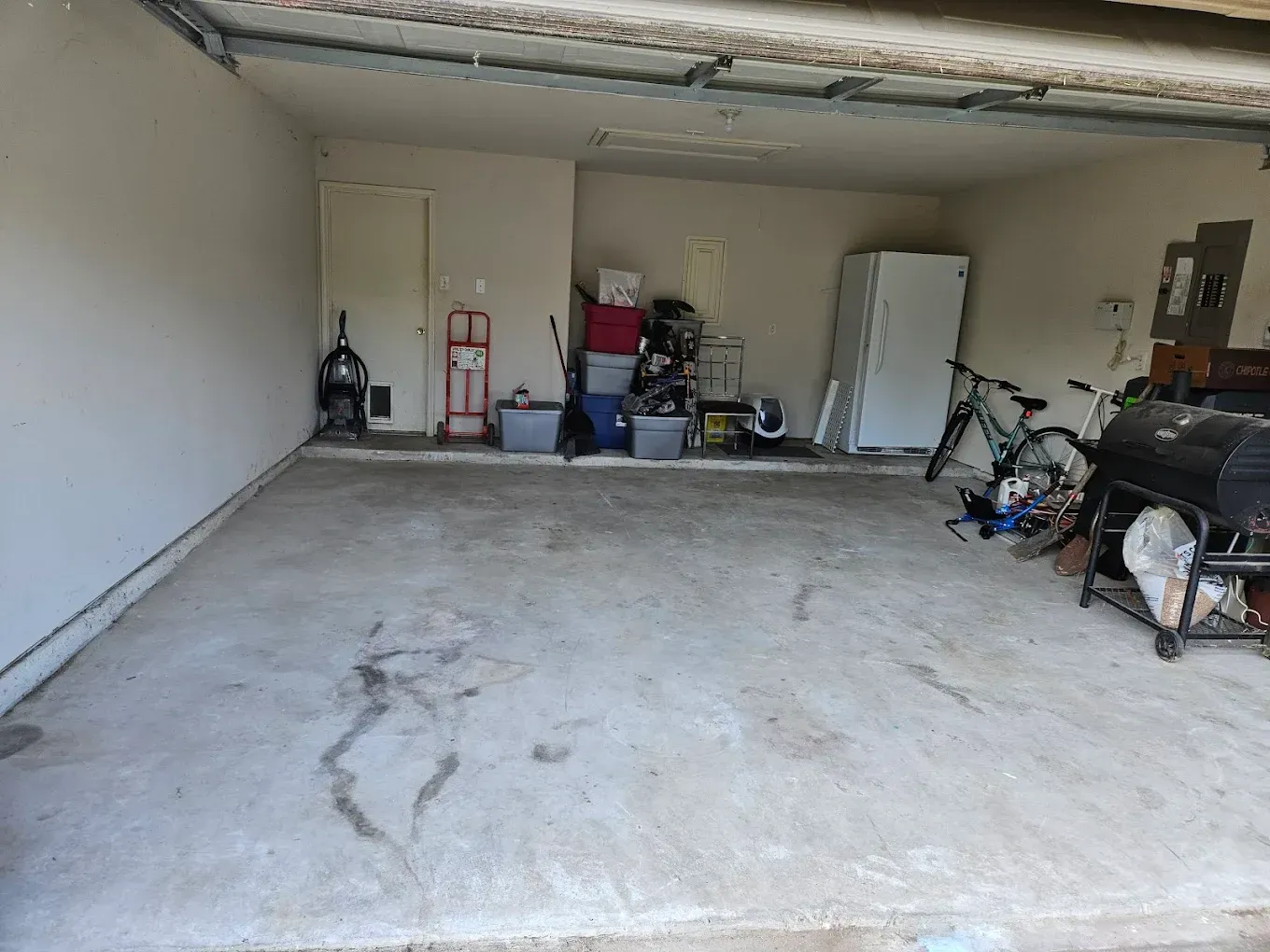 Cluttered garage interior with a concrete floor, shelves, tools, and stored items against the back wall