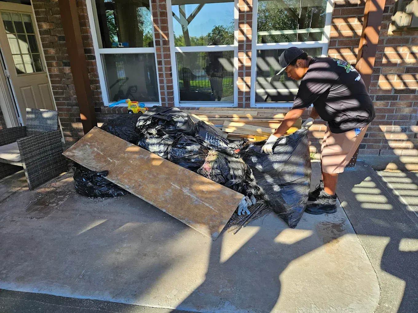 Person clearing debris beside a house porch, with a broken board and pile of roof shingles.