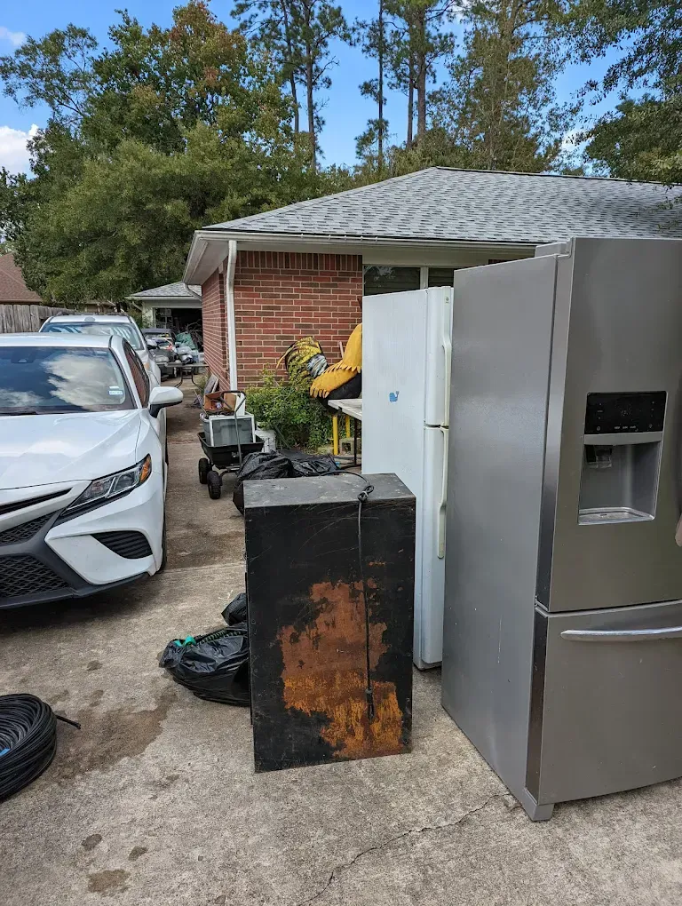 Driveway with a white car, large silver refrigerator, and rusted black metal cabinet near a brick garage.