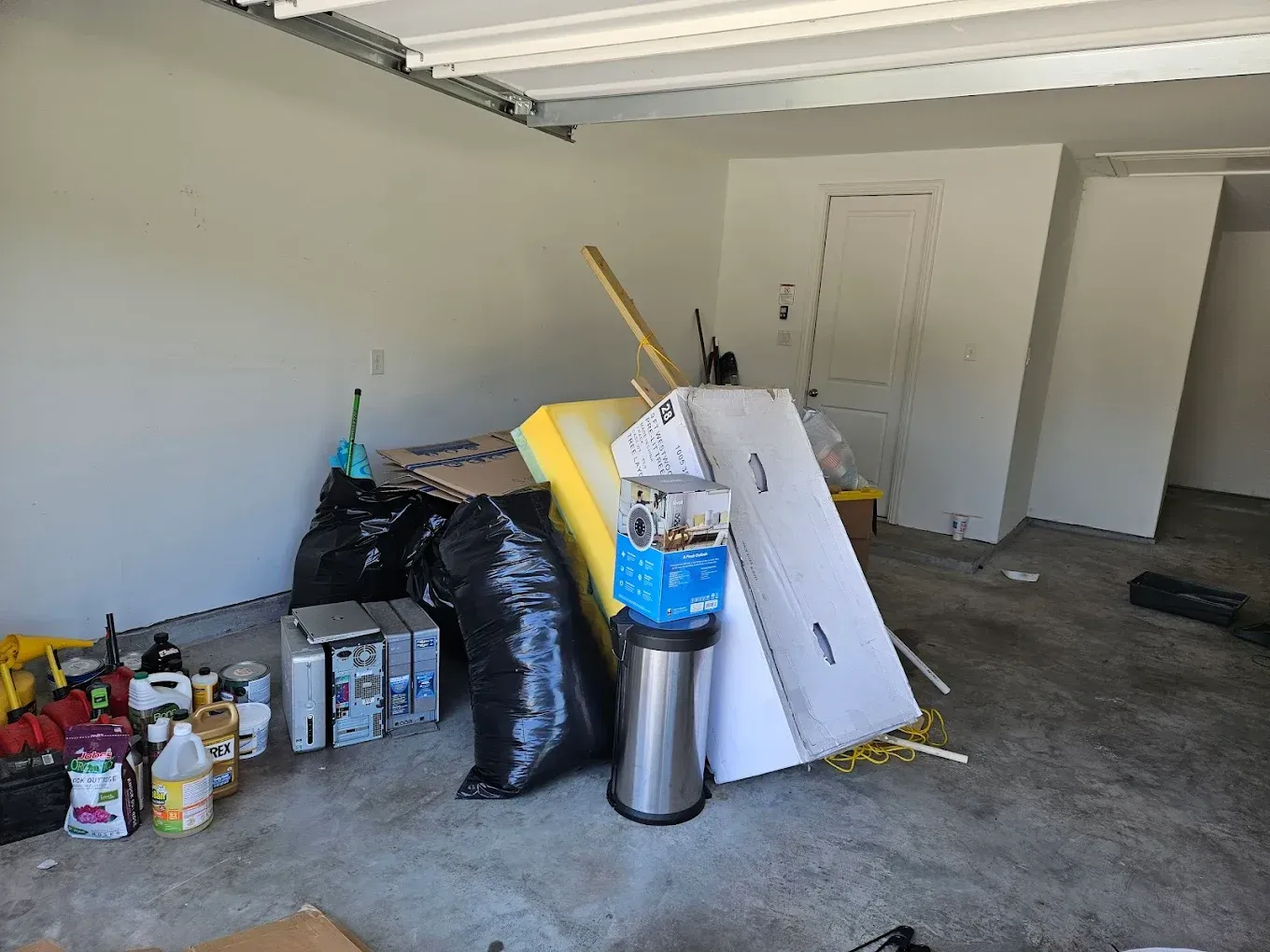 Cluttered garage with trash bags, paint cans, a blue container, and boards leaning against the wall.