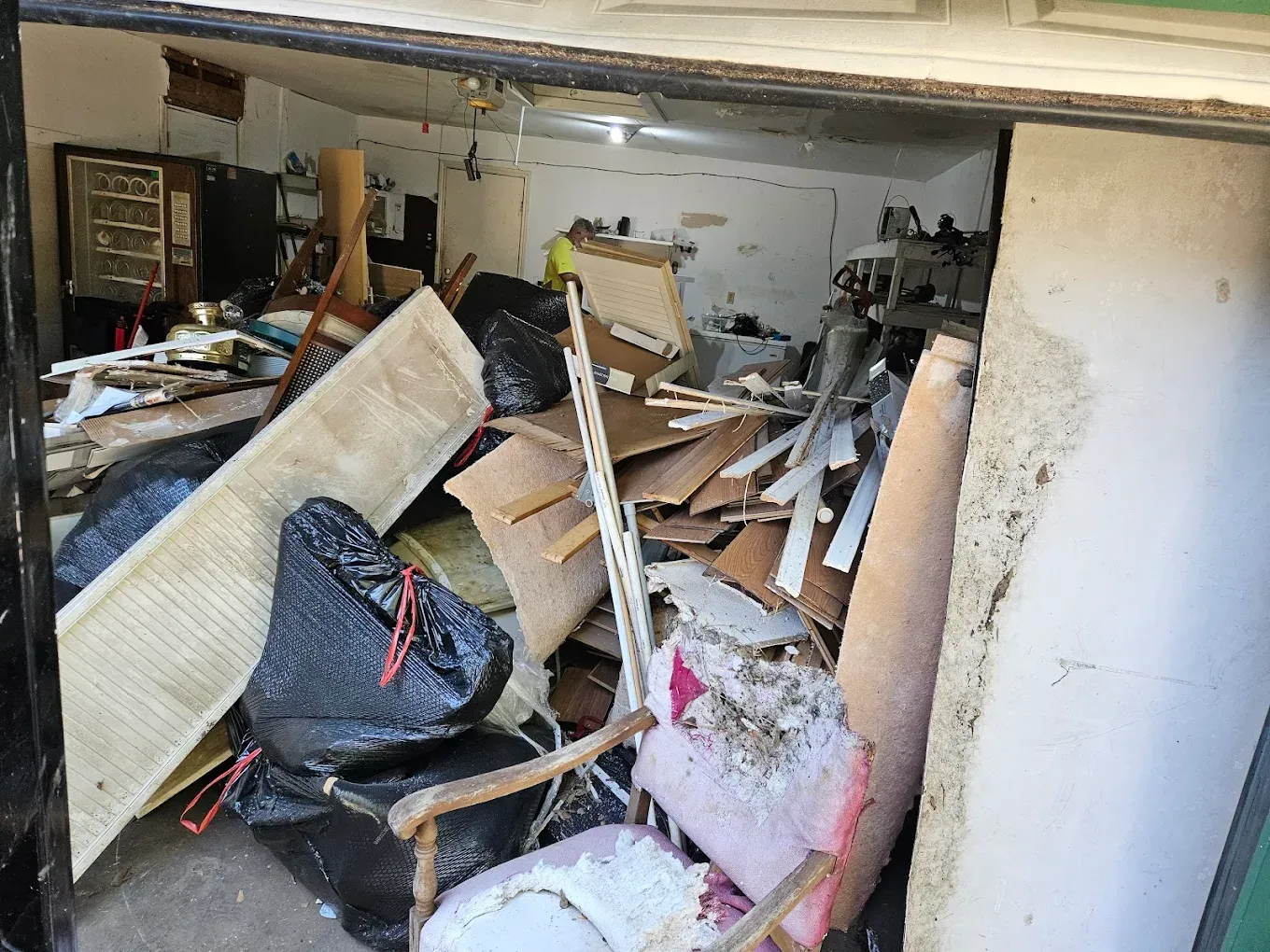 Cluttered garage filled with broken furniture, boards, and trash under a low ceiling
