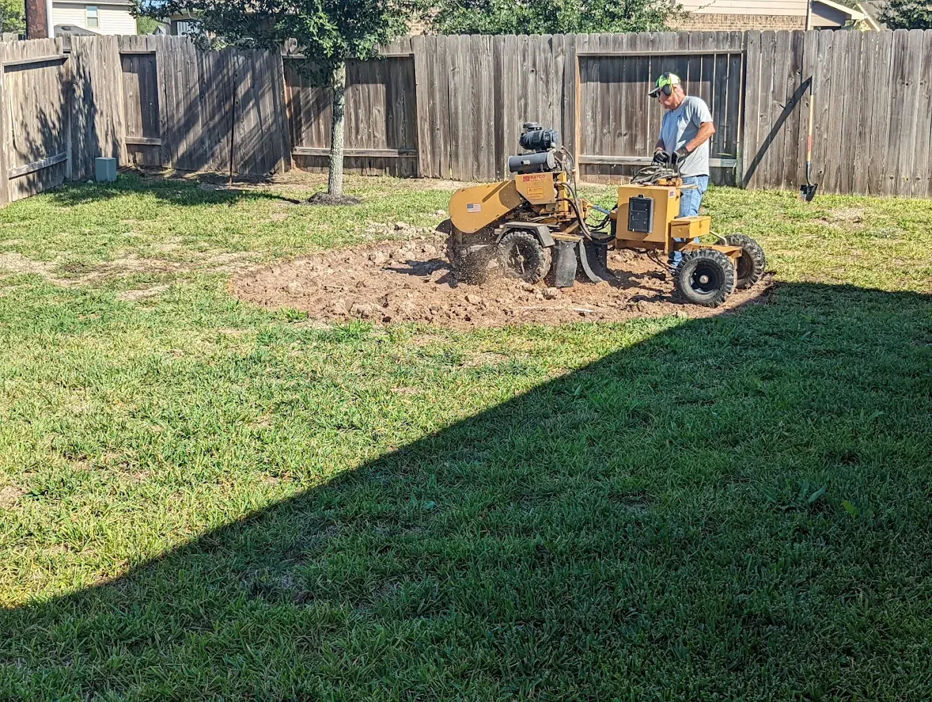Two workers operate a small tractor in a fenced backyard, digging up dirt beside a lawn.
