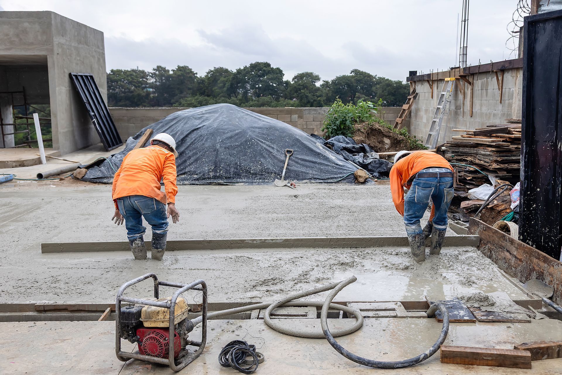 Two workers in orange jackets finishing a concrete slab at a construction site.