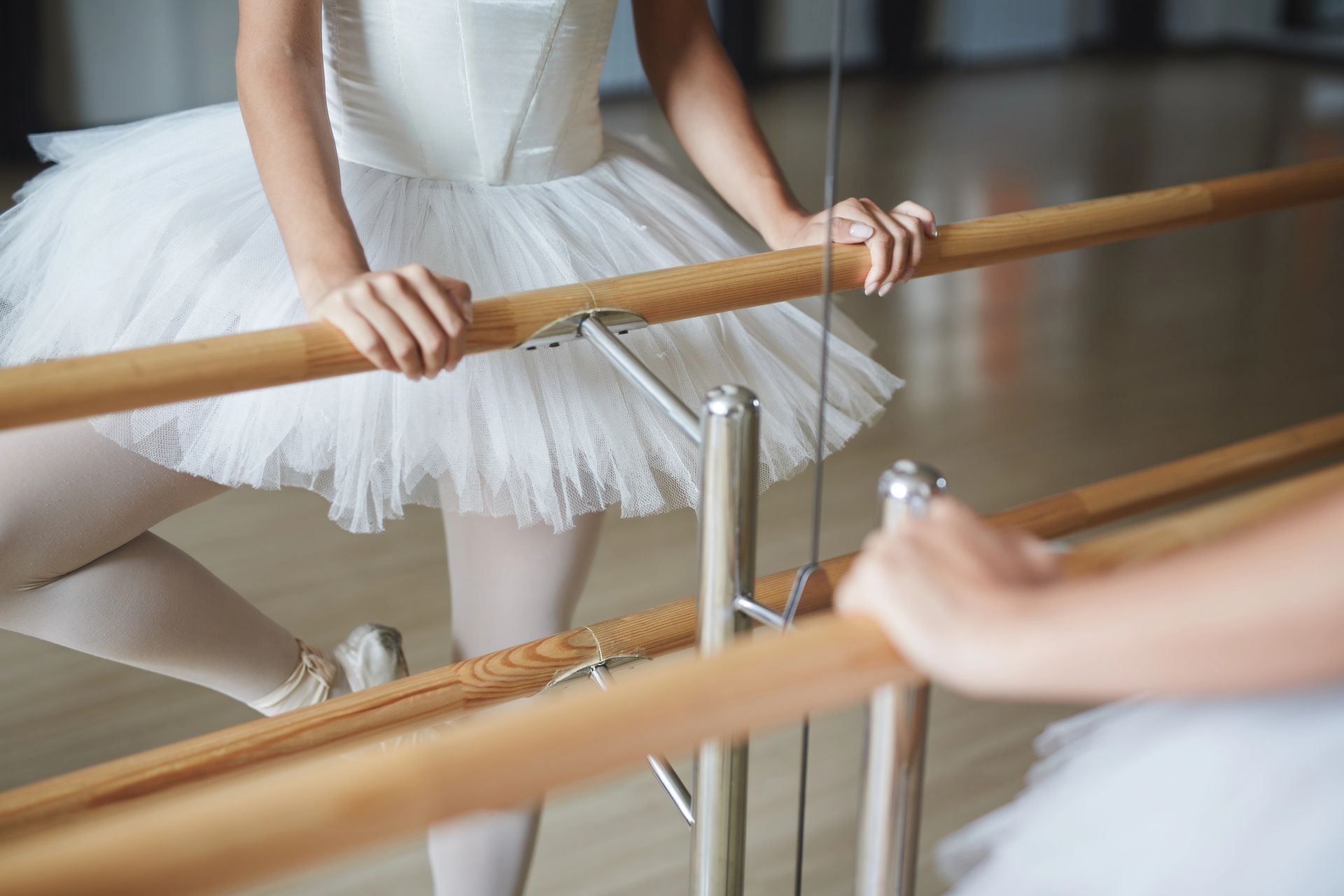 Ballerina in tutù bianco e scarpette da punta che regge la sbarra, mentre si esercita in una sala da ballo con riflessi nello specchio.