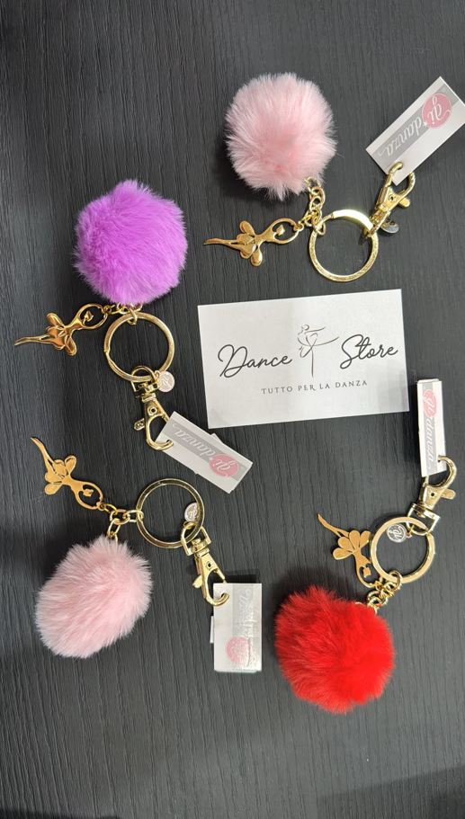 Cinque portachiavi colorati con pompon e dettagli dorati, su una superficie scura, vicino a un biglietto.