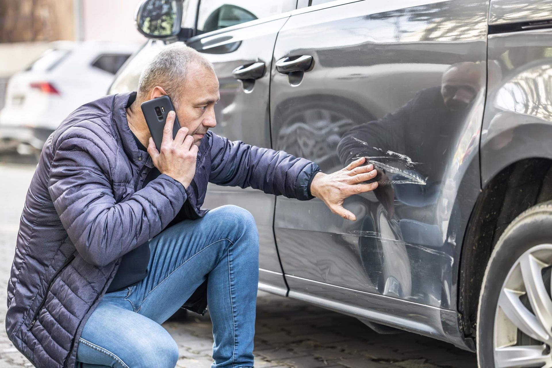 Man on phone examines dented gray car door, outdoors.