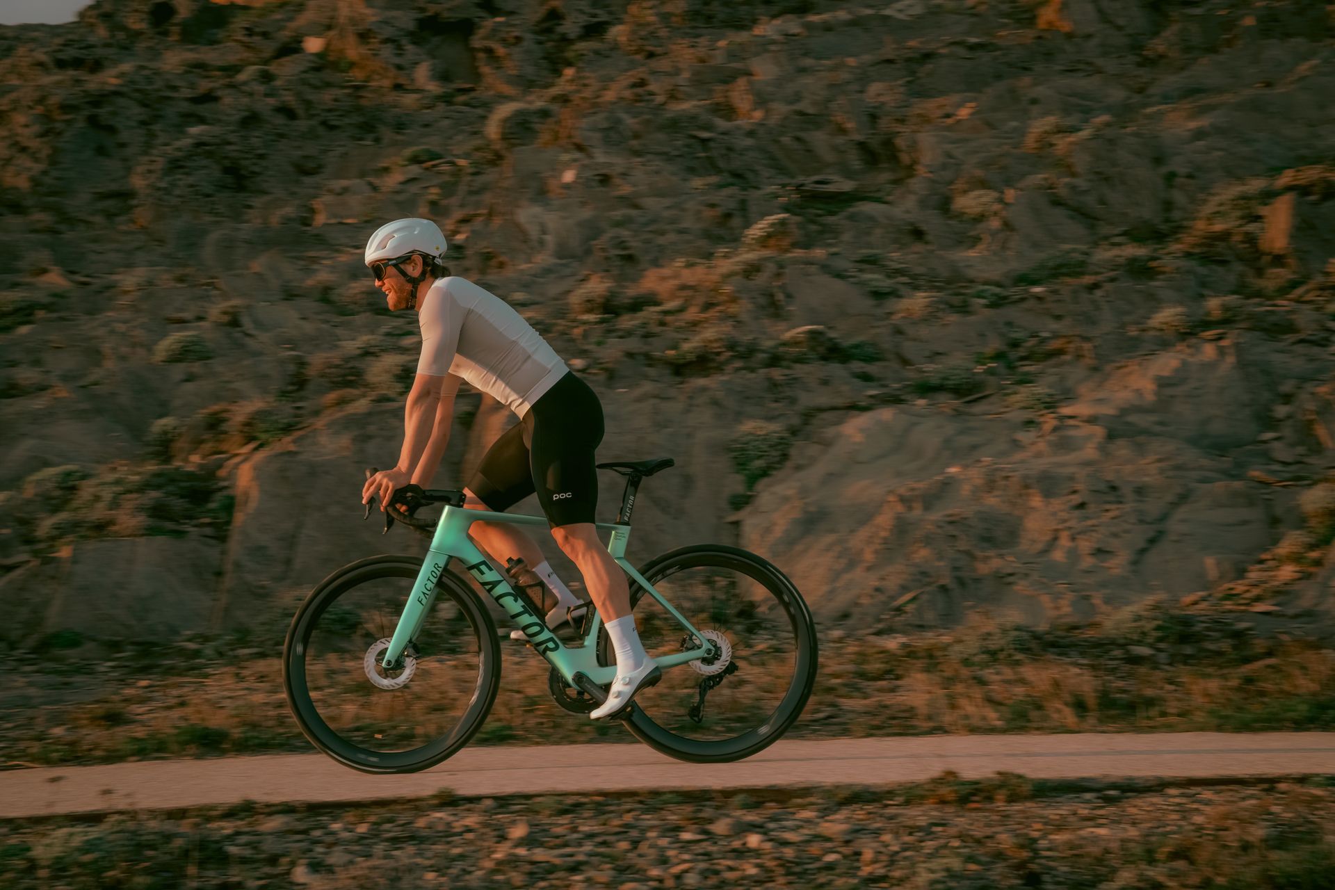 Cyclist in light gear riding a turquoise bike on a dirt path, rocky background, setting sun.