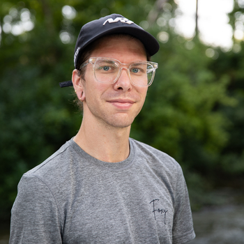 A man wearing glasses and a hat is standing in front of a river.