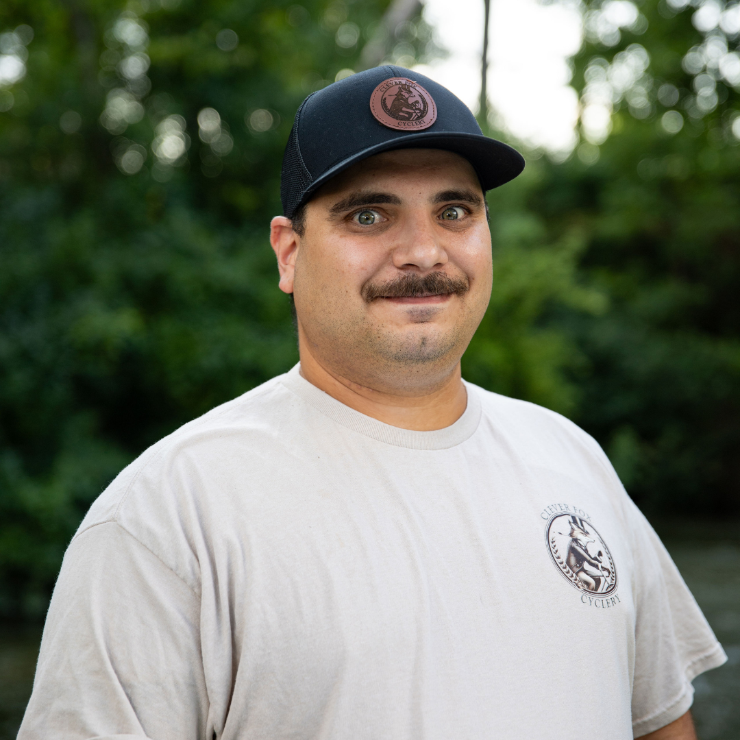 Man wearing a black cap and white T-shirt outdoors, standing in front of green foliage.