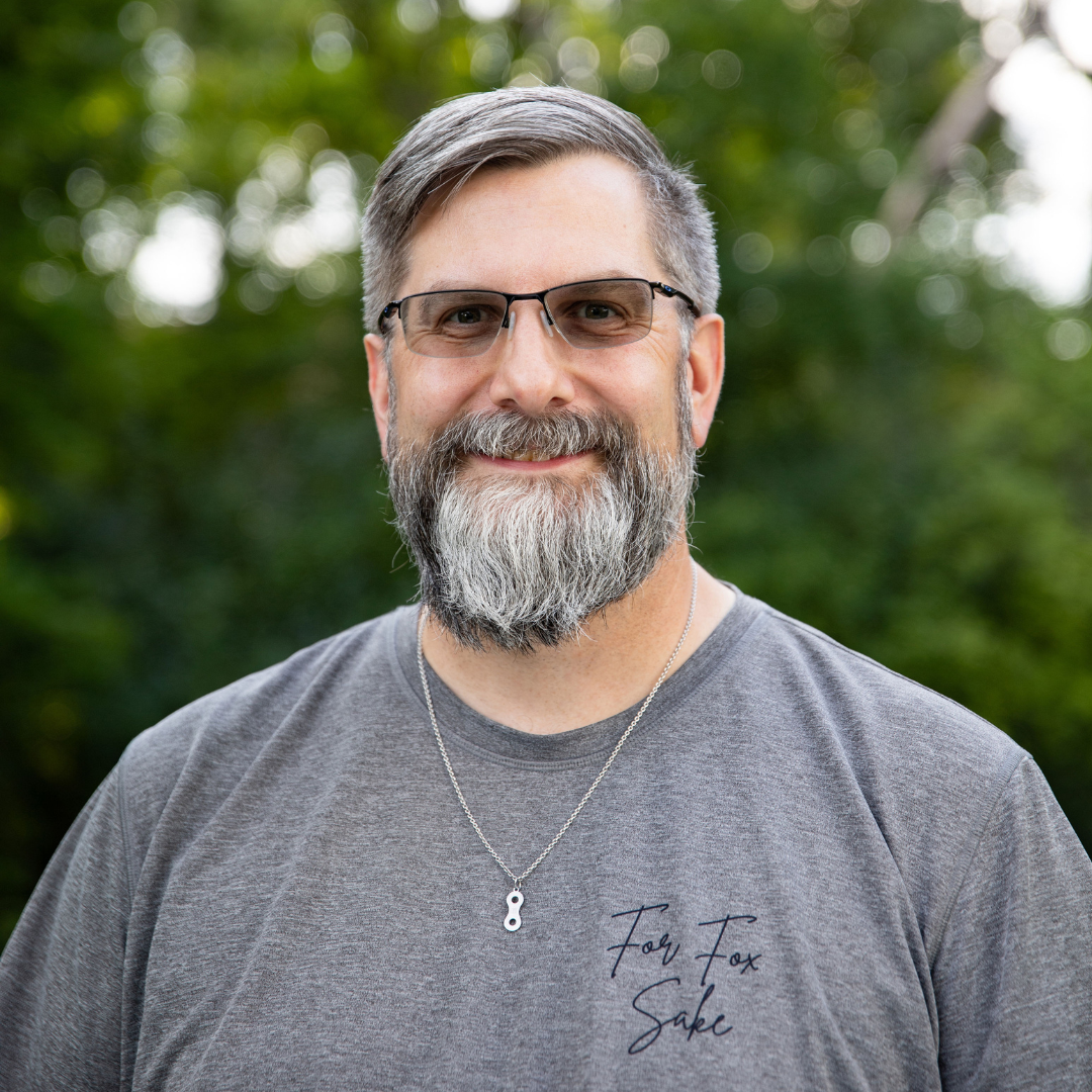Smiling man with glasses and gray beard wearing a gray T-shirt outdoors with blurred greenery in the background
