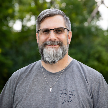 A man with a beard and glasses is wearing a gray shirt and a necklace.