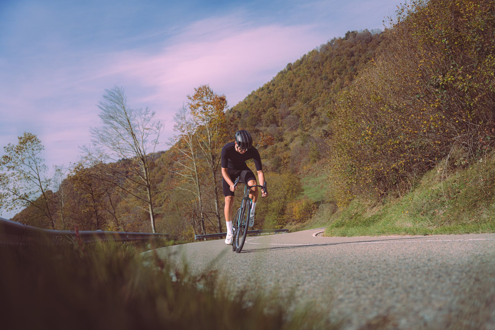 Cyclist riding uphill on a rural road beside a grassy hillside under a pink sky