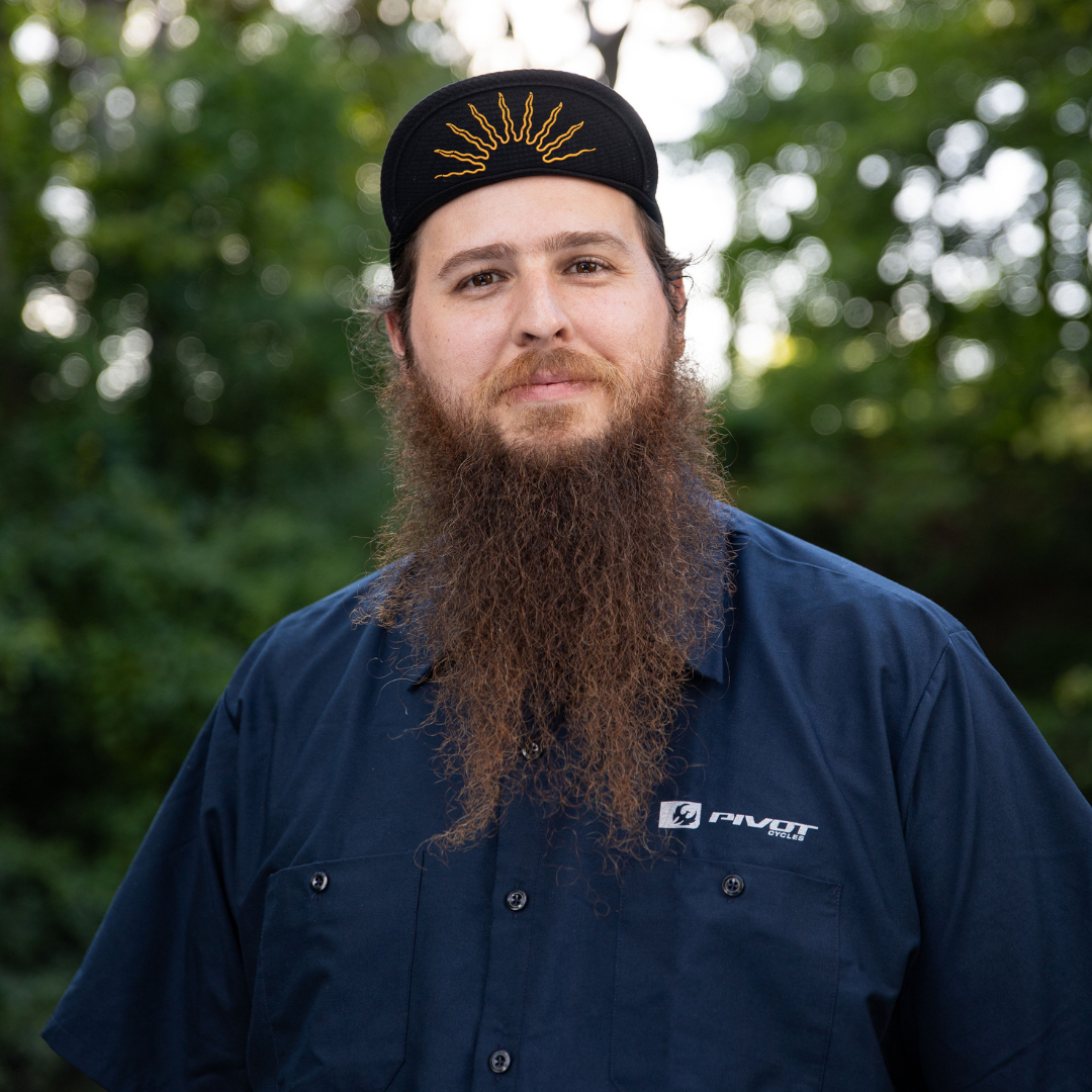 Bearded man in a navy shirt and black cap standing outdoors with blurred greenery behind him
