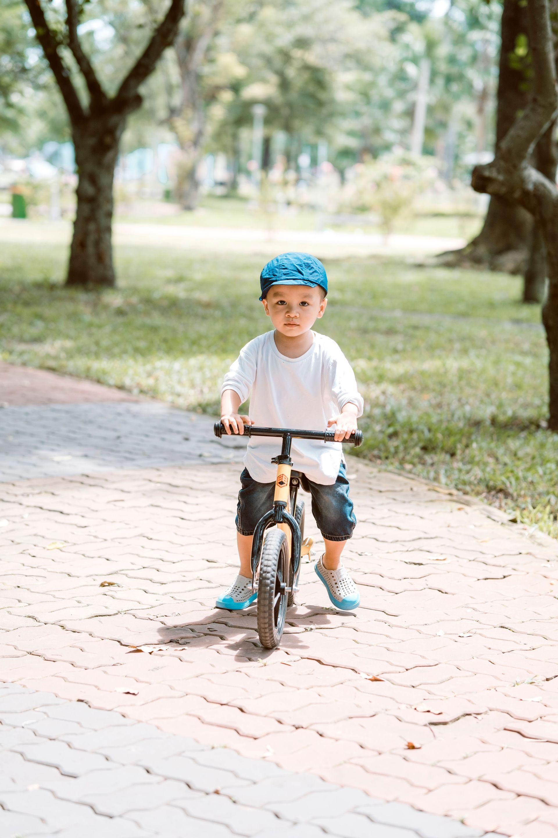 Child riding a bicycle on a tree-lined park path, wearing a blue helmet and white shirt.
