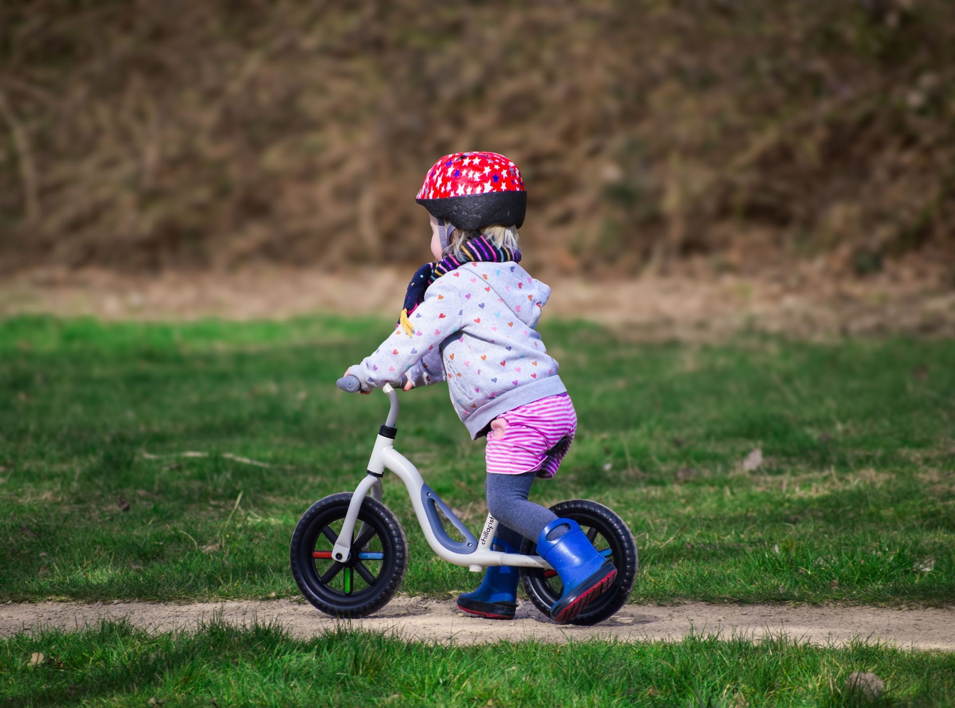 Child riding a balance bike on a grassy path, wearing a red helmet and gray jacket