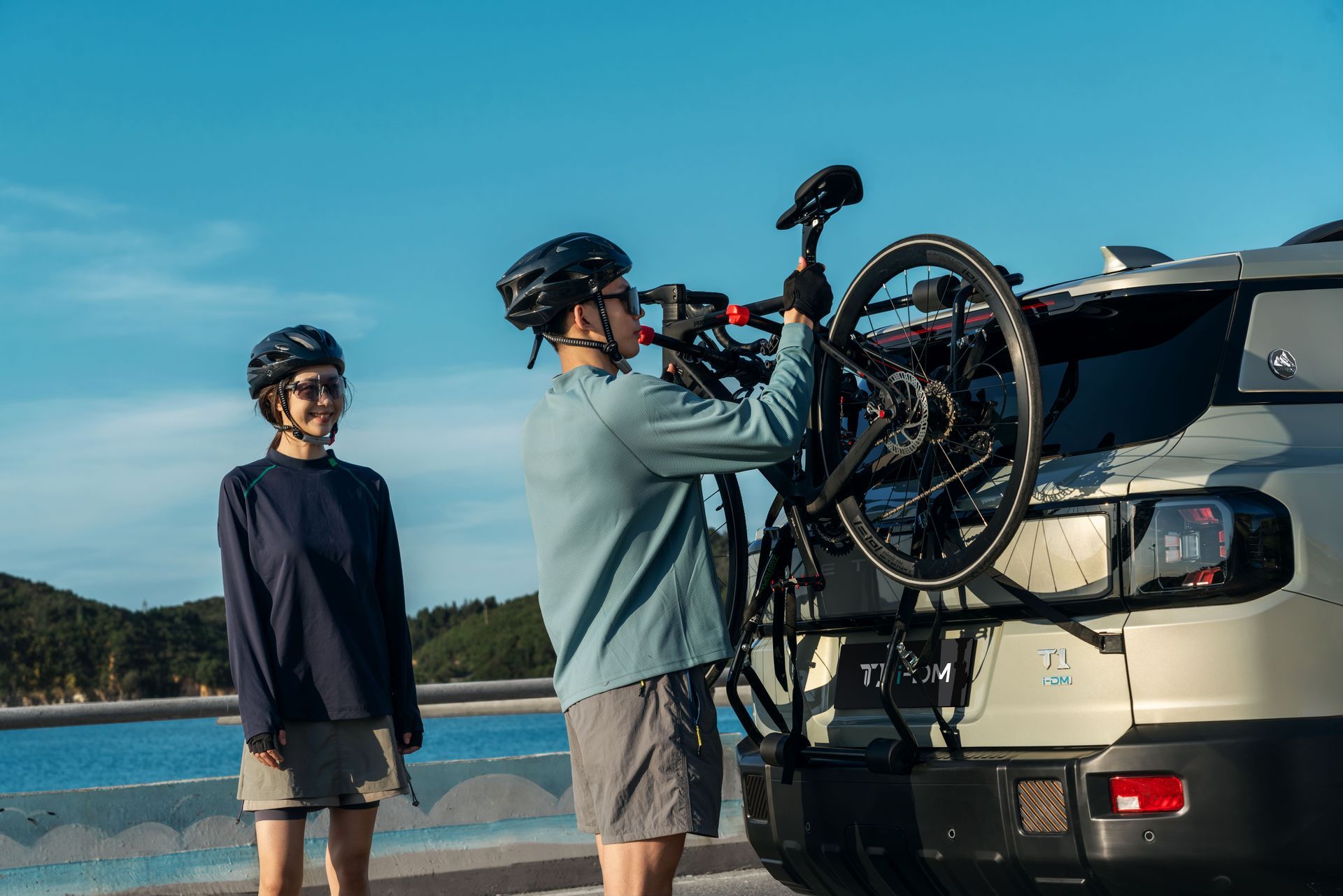 Two cyclists load bikes onto an SUV by a lakeside road.
