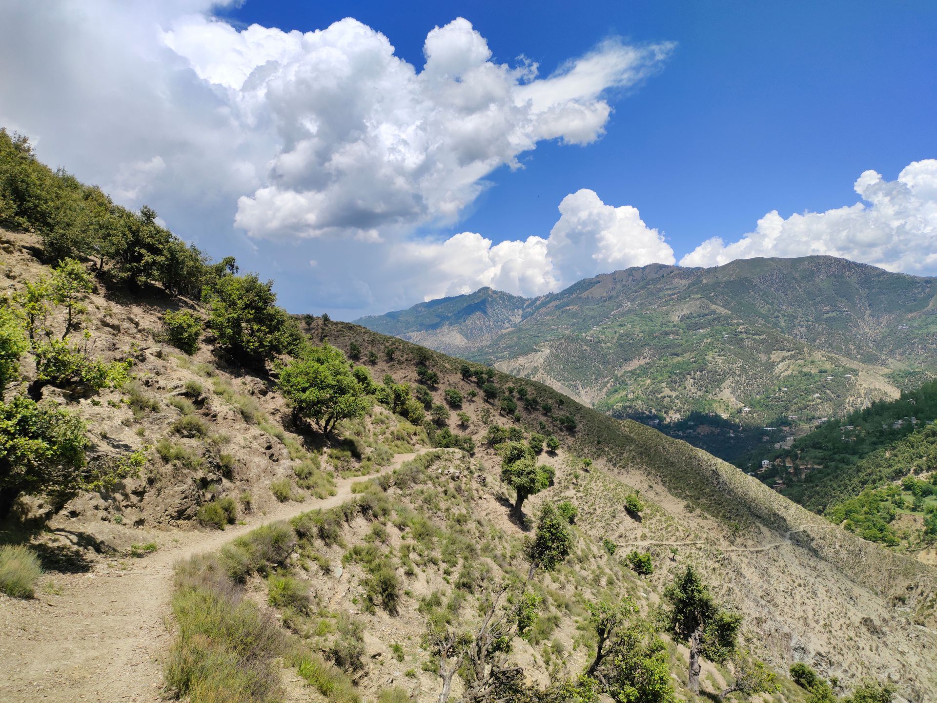 Mountain trail winding through dry hills under a bright blue sky with clouds and distant peaks