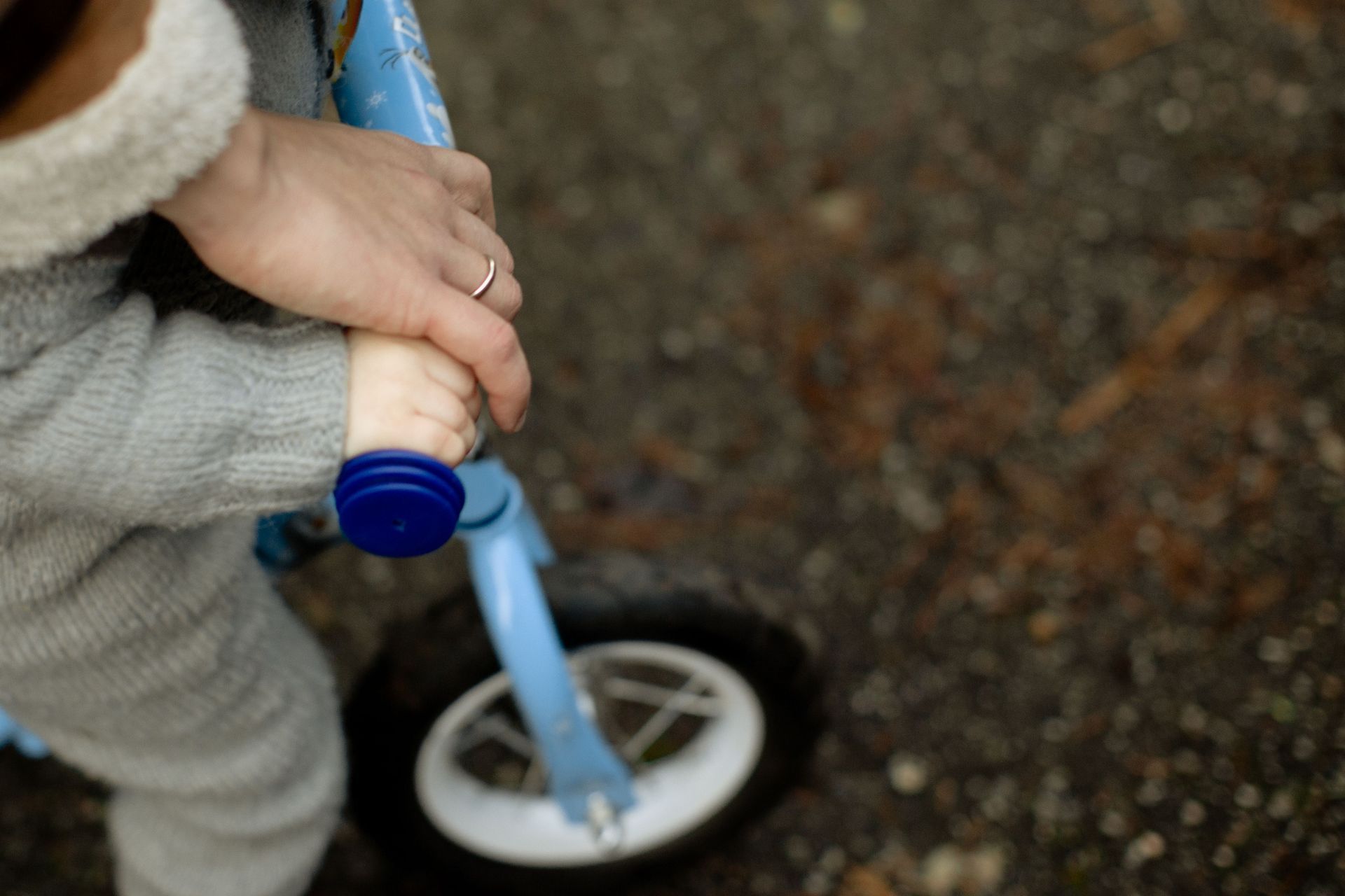 Child holding a blue tricycle wheel on a gravel path, with an adult hand assisting.