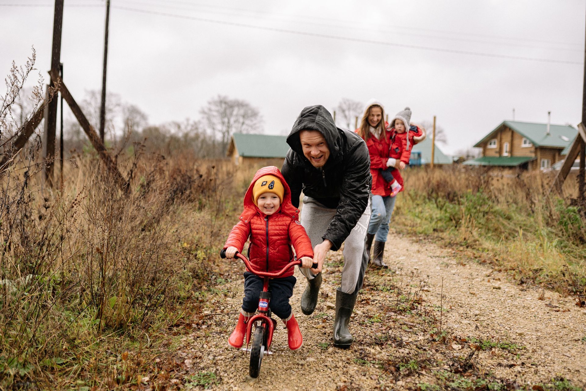 Adults and child in red rain gear riding and pushing a tricycle on a muddy country path