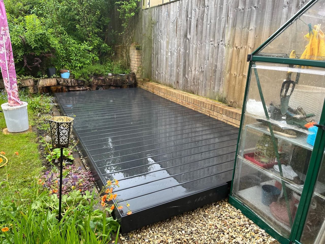 Dark grey composite deck running the length of a narrow garden beside a timber fence and a greenhouse