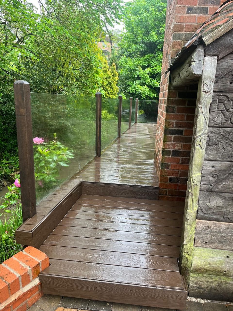 Brown composite deck along a narrow side return beside a brick house, with a glass balustrade on timber posts and a step down