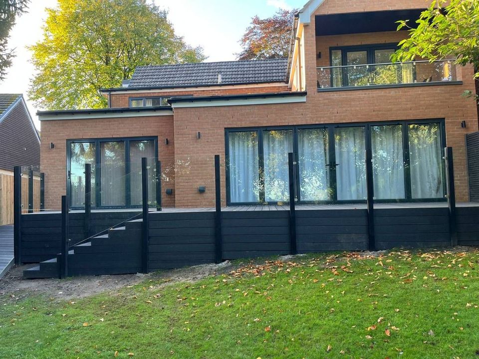 Long raised black composite deck with a glass balustrade and steps down to the lawn along the back of a brick house