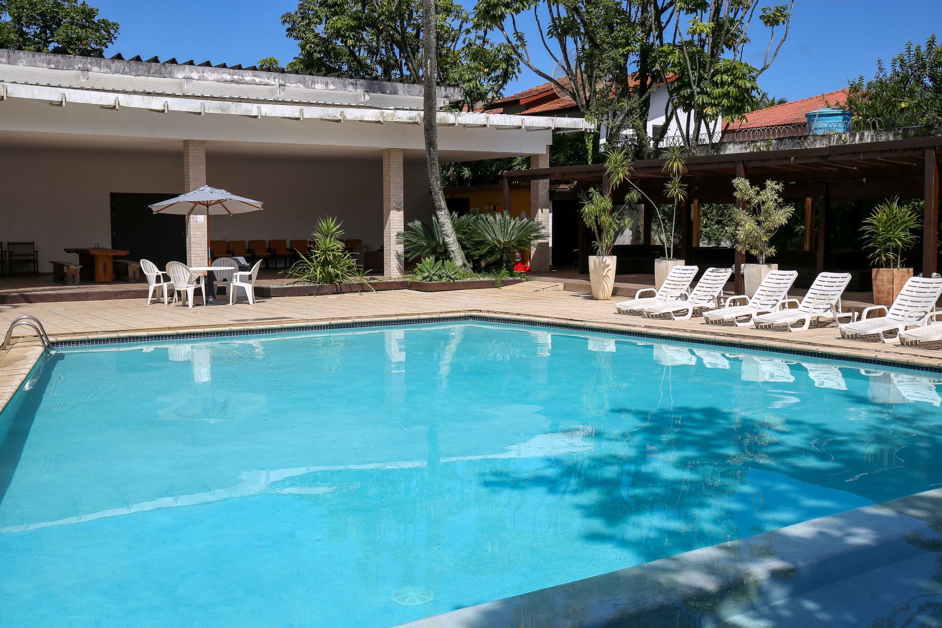 Piscina com água azul, espreguiçadeiras brancas, guarda-sol e área de pátio coberta sob um céu ensolarado.