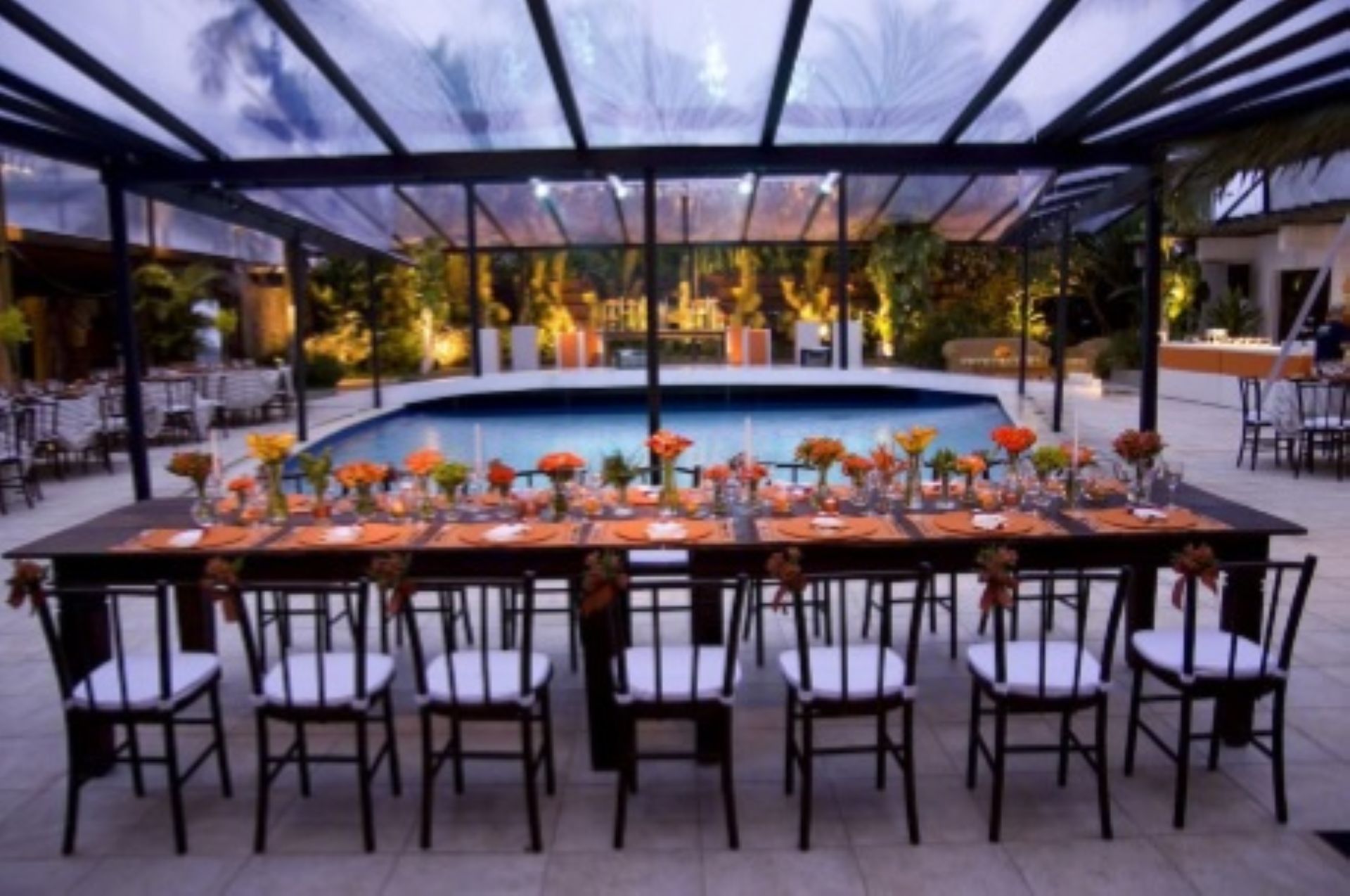 Mesa comprida posta para uma festa com piscina ao fundo, sob um teto transparente.