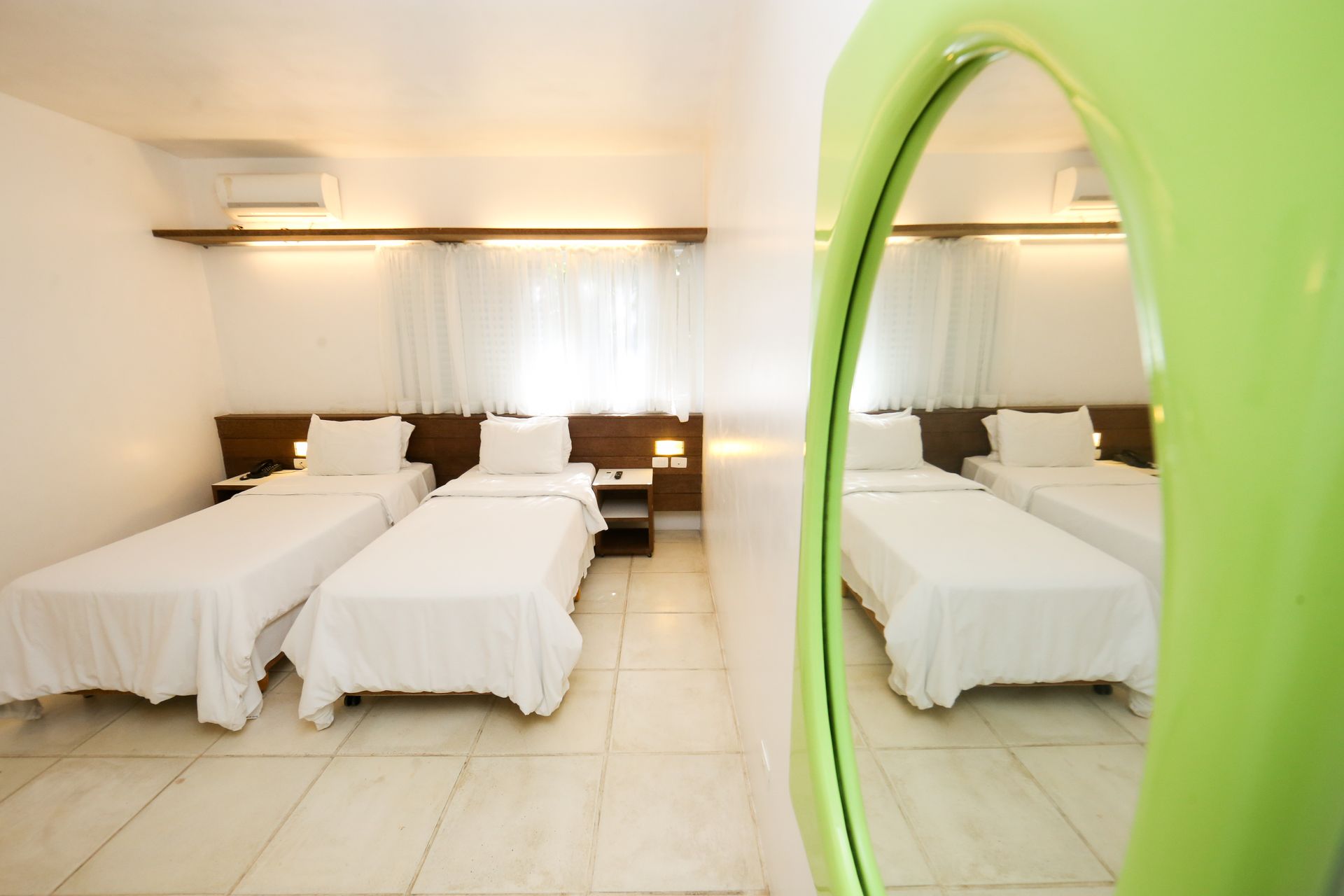 Quarto de hotel luminoso com duas camas, refletidas em um espelho com moldura verde. Paredes brancas, piso de azulejo e decoração simples.
