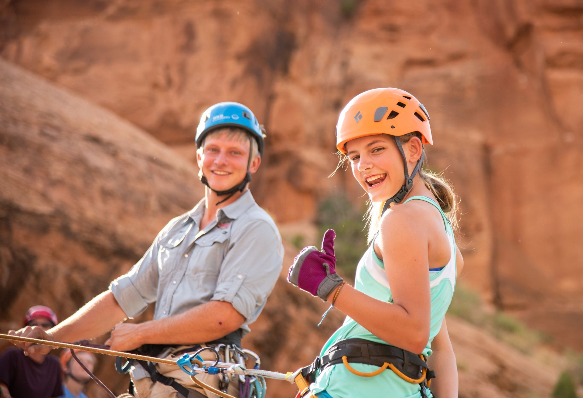 Man and girl in helmets smiling outdoors, holding ropes near red rock. Girl gives thumbs up.
