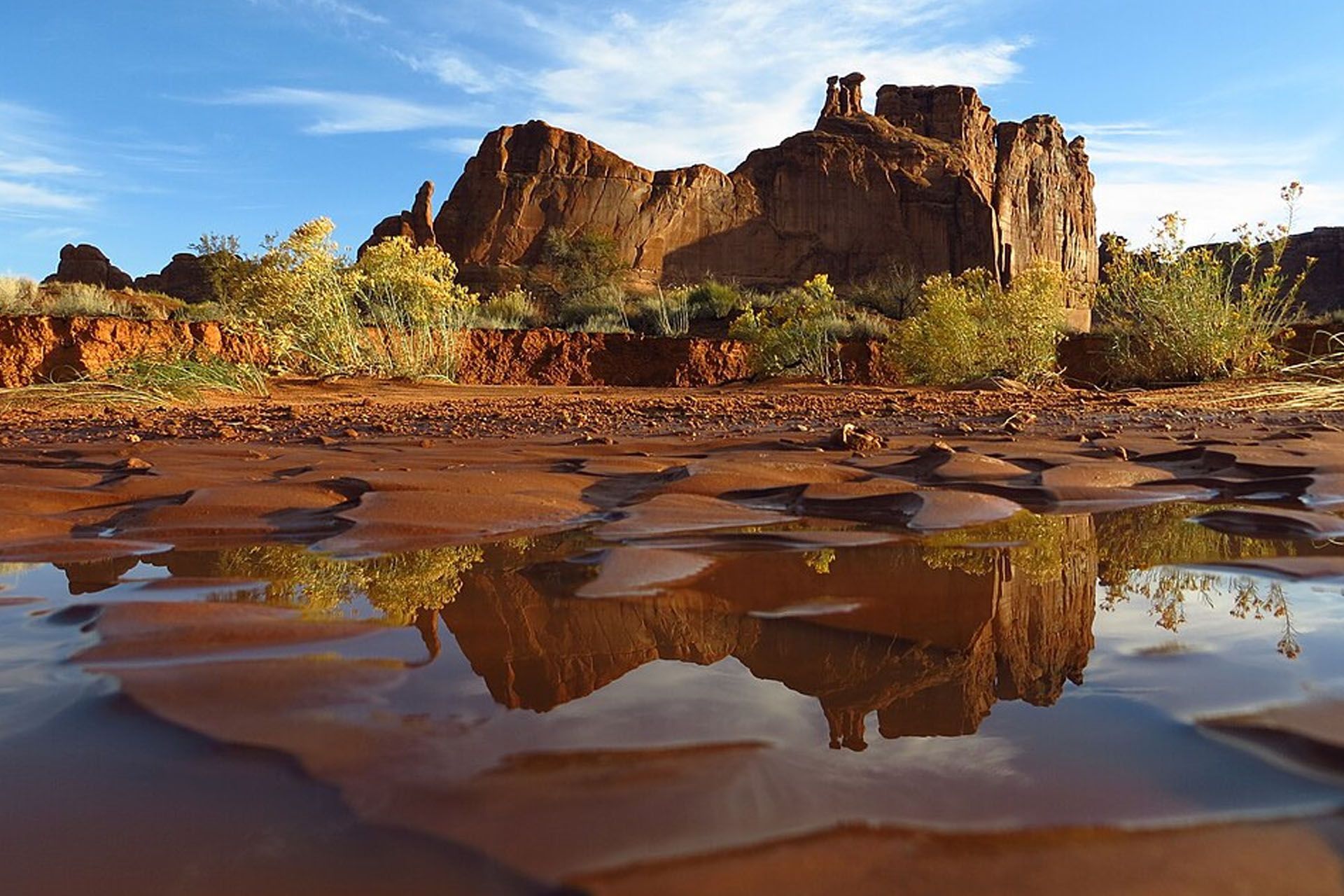 Red rock formation reflected in a shallow pool of water, under a blue sky.