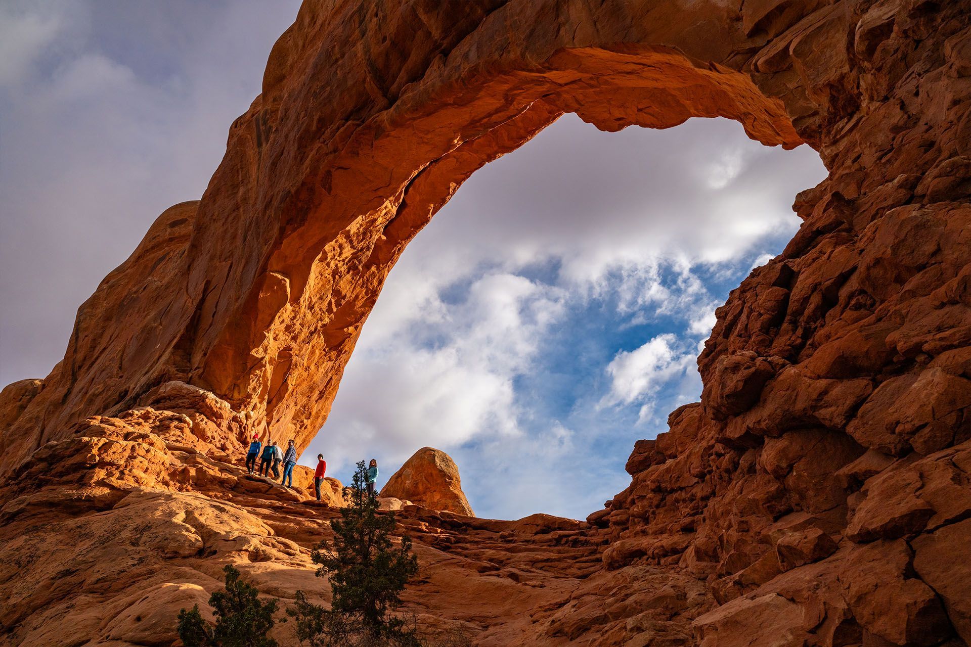 Large sandstone archway with people standing on the rocky top, blue sky and clouds visible through the opening.