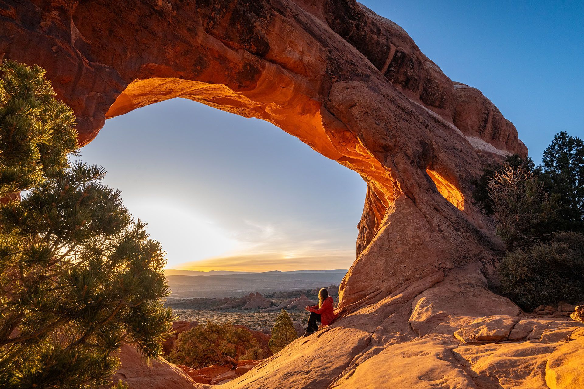 A person sits under a large arch with the early morning sun glowing across the rocks.