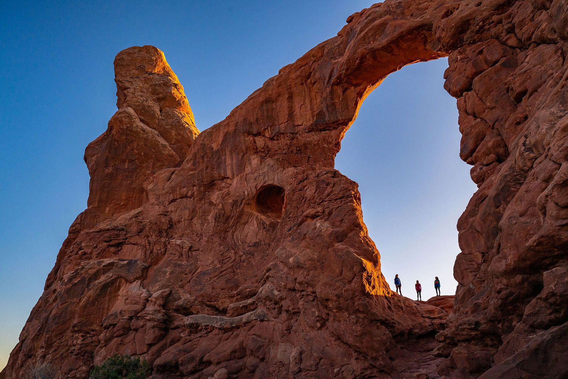 Red rock archway framing silhouetted hikers against a blue sky.