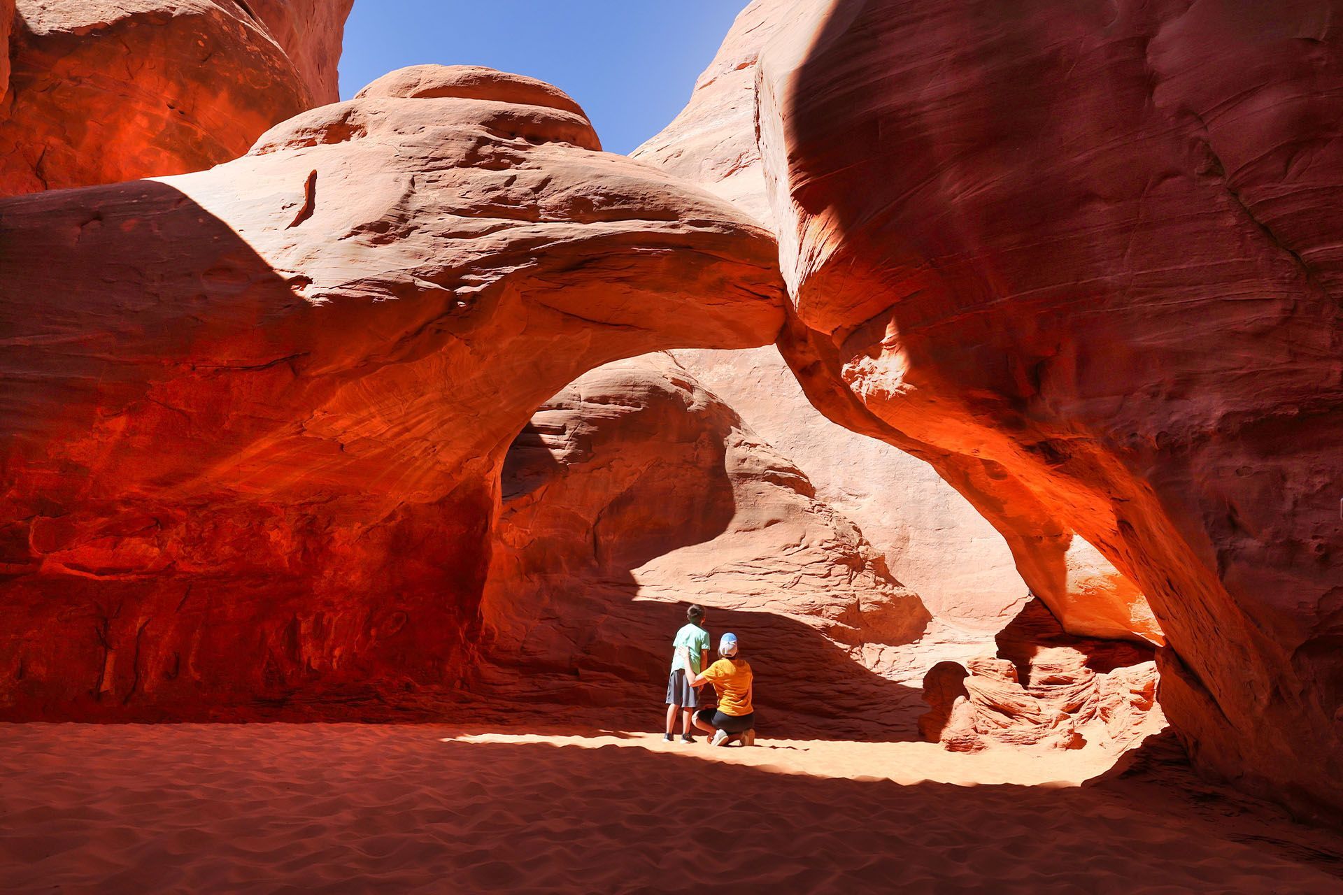 Sand Dune Arch with two people standing inside, bright sunlight casting shadows.