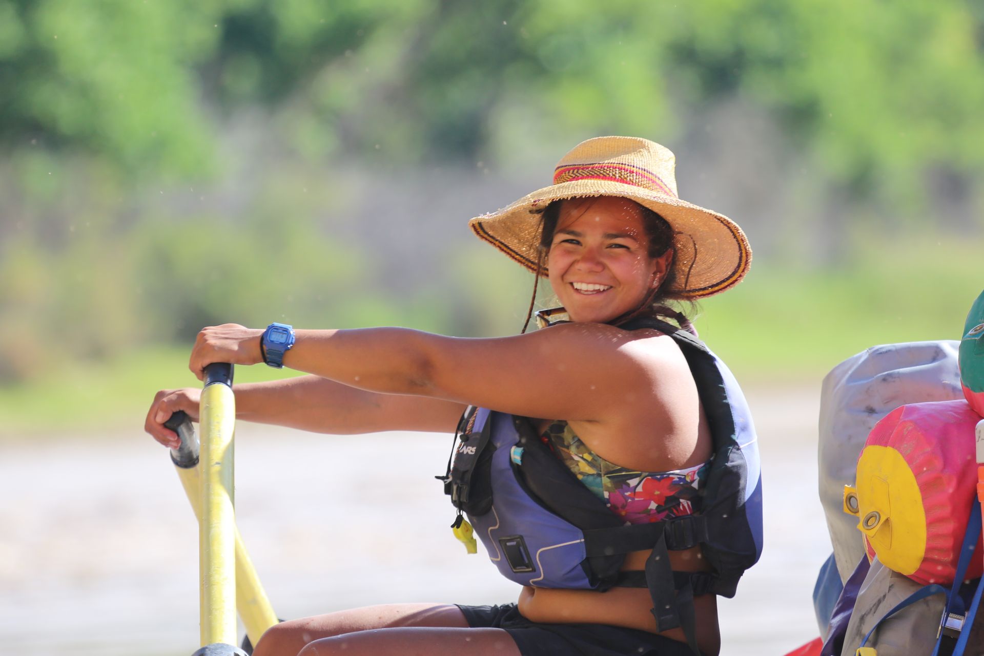Woman in straw hat and life vest smiles, holding oars on a raft; blurred river background.