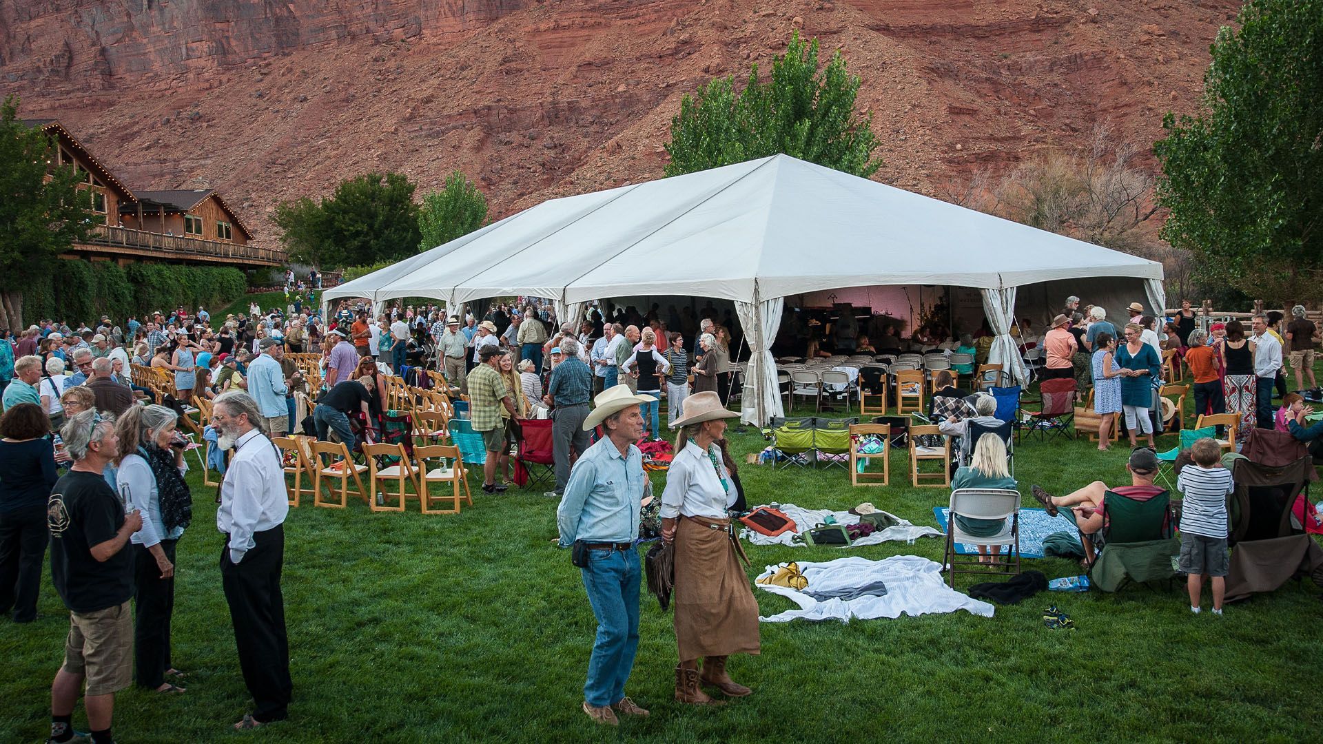 Large outdoor event under a white tent in front of red rock cliffs. People mingle on the grass.