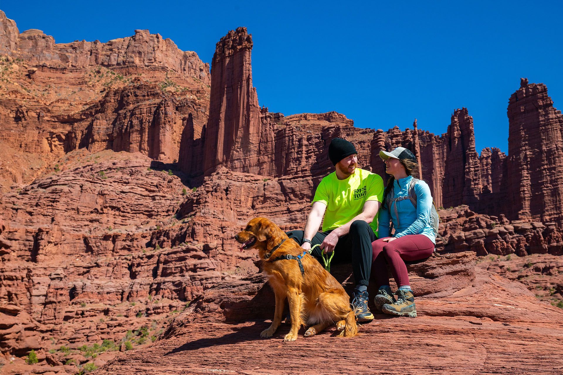 Two hikers hold their dog on a leash with Fisher Towers standing tall behind them.