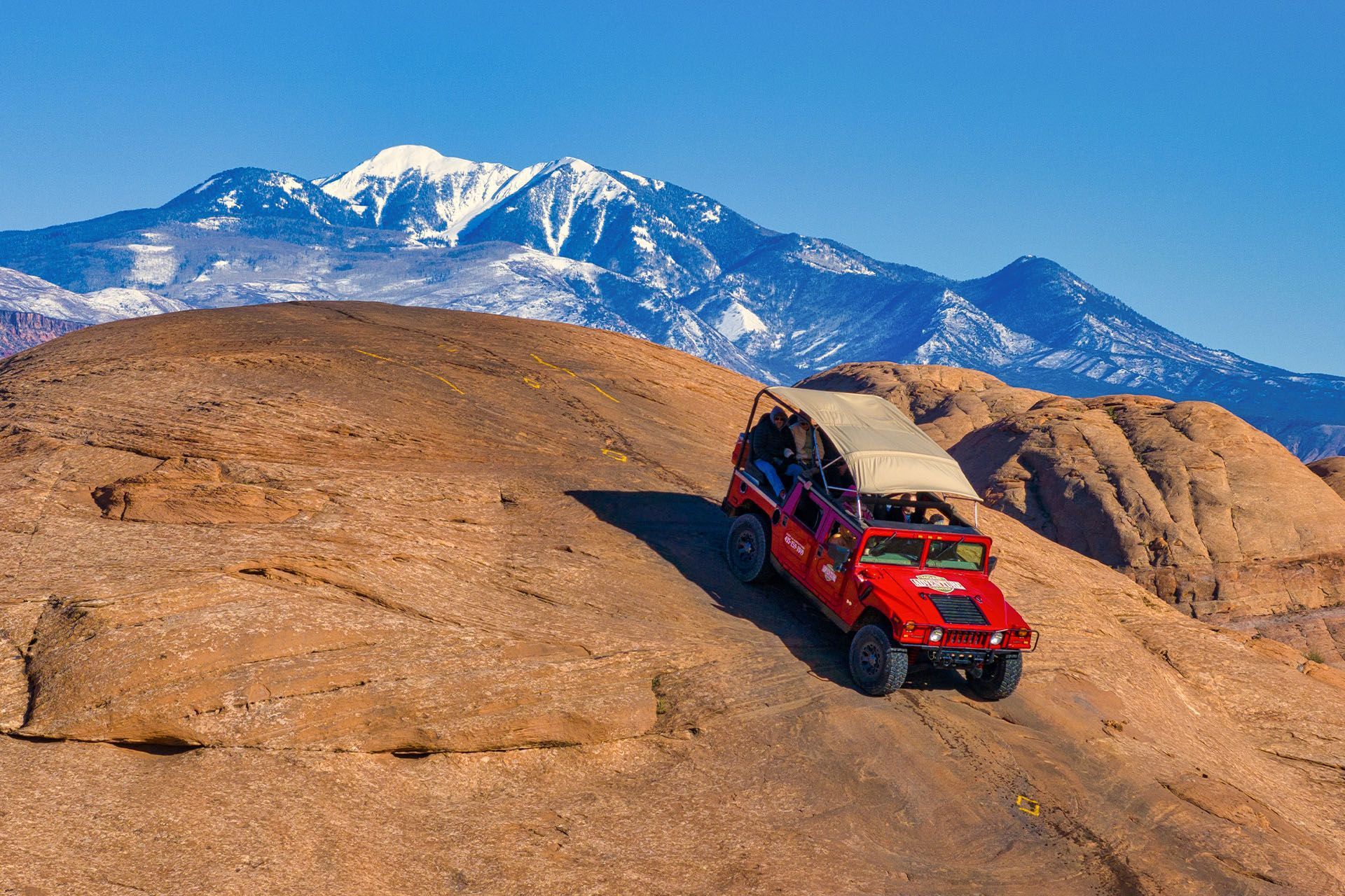 Red off-road vehicle climbing a rocky hill with snow-capped mountains in the background.