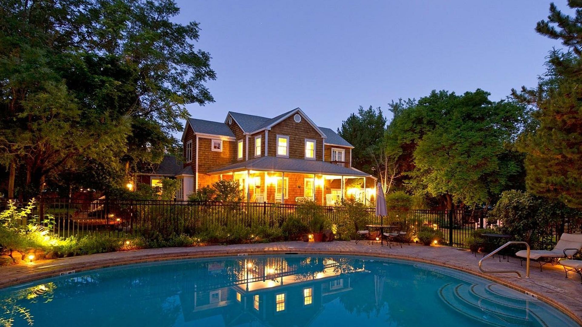 Quaint bed and breakfast with a pool at dusk; warm lights illuminate the porch and trees surrounding the pool.
