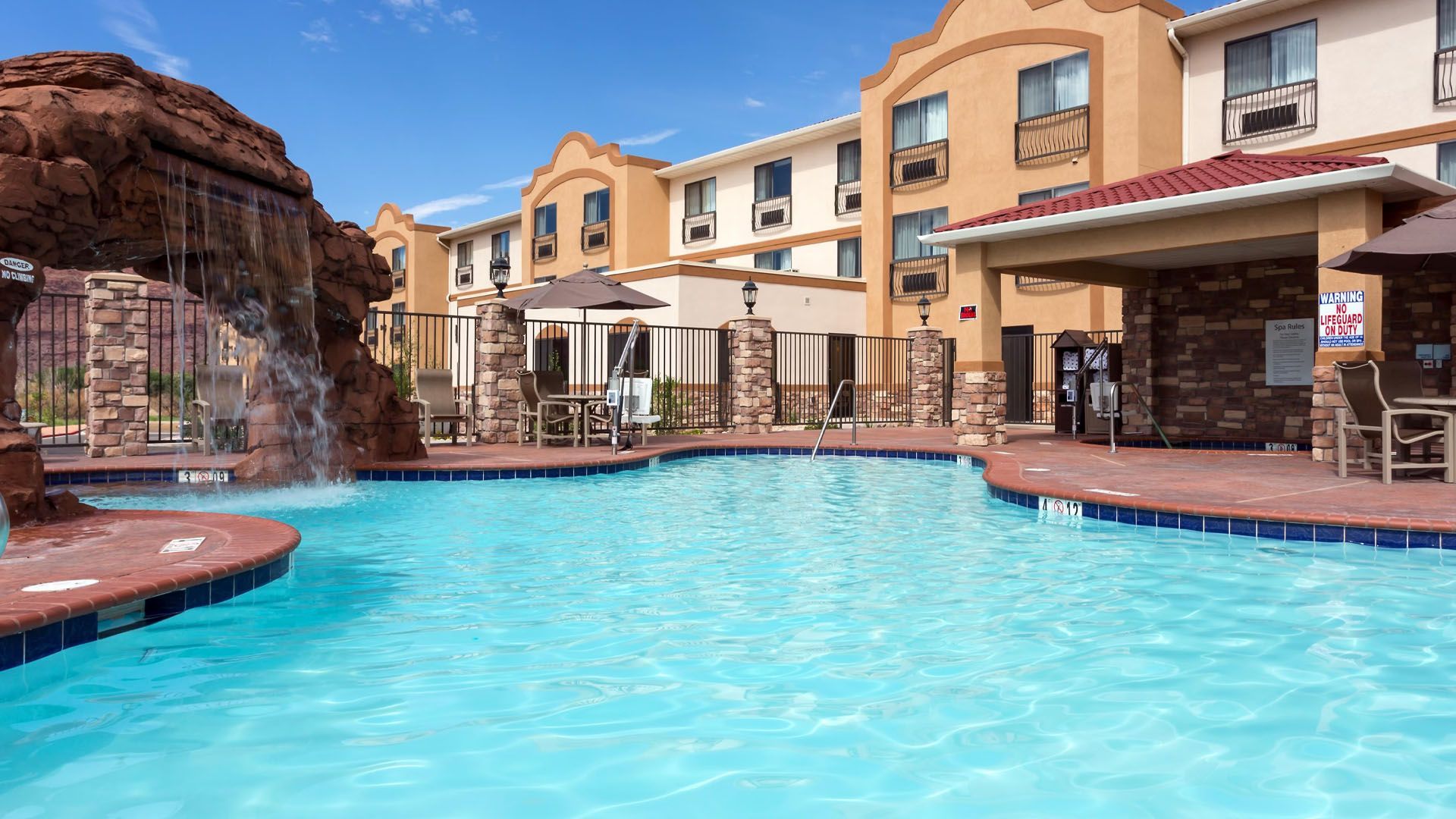 Hotel pool with a waterfall feature and building in the background under a bright sky.
