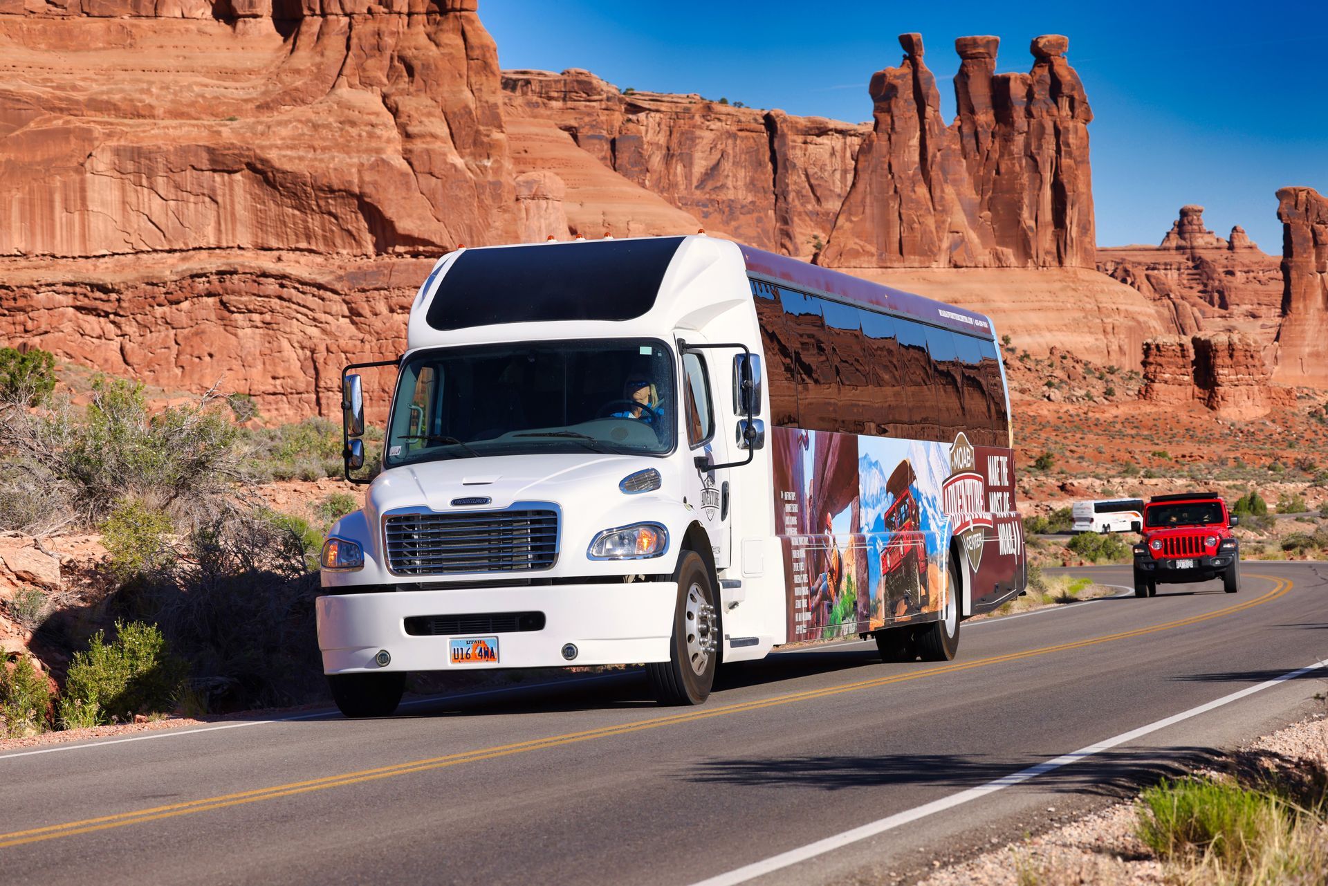 White tour bus on road in front of red rock formations, small red SUV in distance.