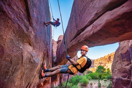A man and woman are rapelling under a stone arch with a canyon below.