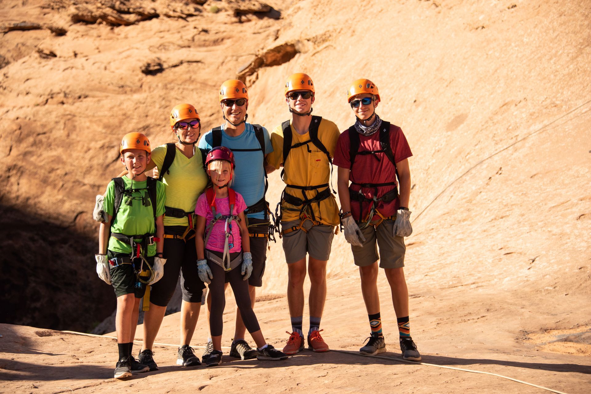 Family of six in climbing gear smiling, posing in front of a rock wall with adults and young children.